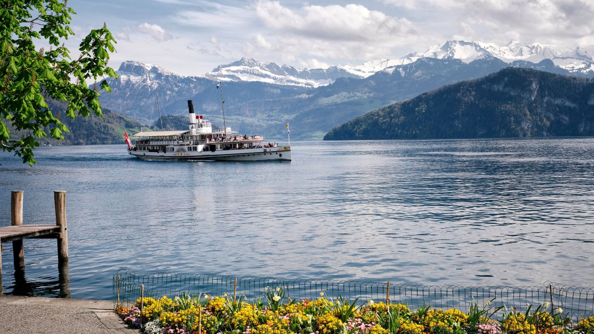 A historic steamboat cruises across Lake Lucerne, with colorful spring flowers in the foreground and snow-capped mountains in the background.