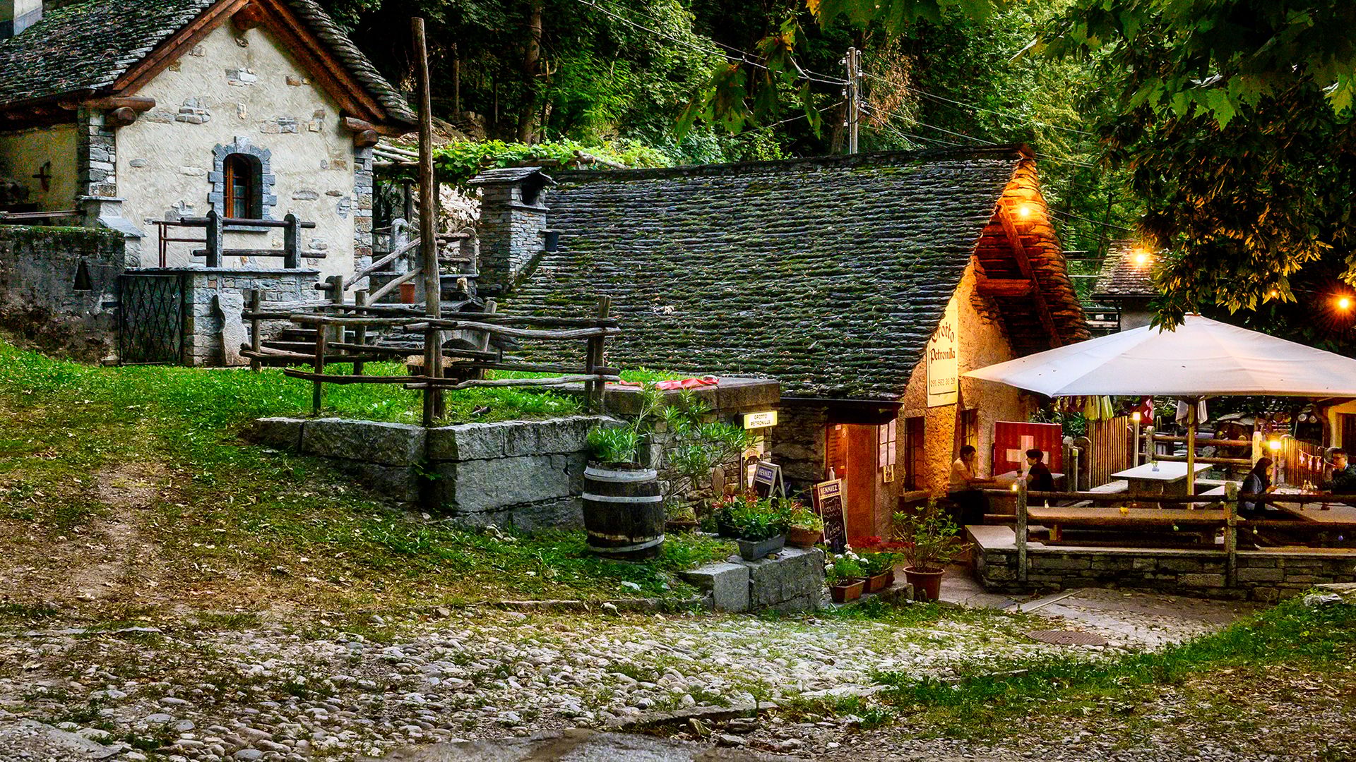Stimmungsvolle Abendaufnahme des Grotto Petronilla in Biasca mit traditionellen Steinhäusern, beleuchteter Terrasse und gemütlicher Atmosphäre im Grünen.