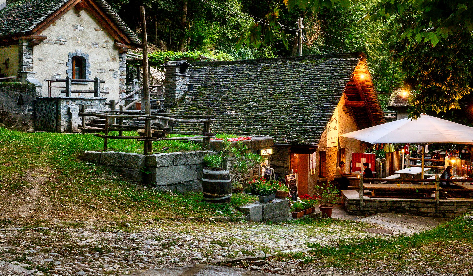 Stimmungsvolle Abendaufnahme des Grotto Petronilla in Biasca mit traditionellen Steinhäusern, beleuchteter Terrasse und gemütlicher Atmosphäre im Grünen.