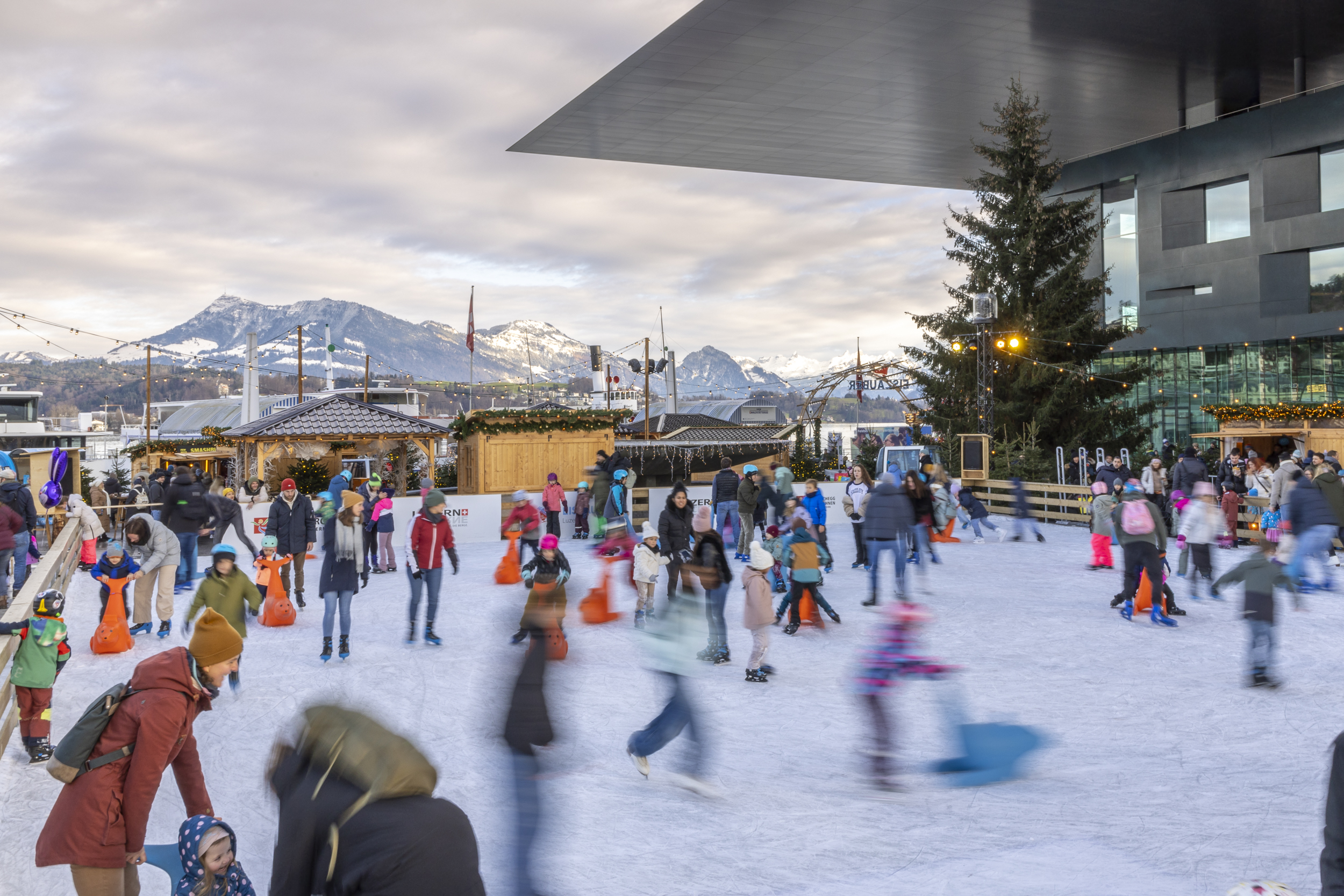 Grosses Schlittschuhfeld mit Menschen und Holzhäuschen rundum mit Weihnachtsdeko