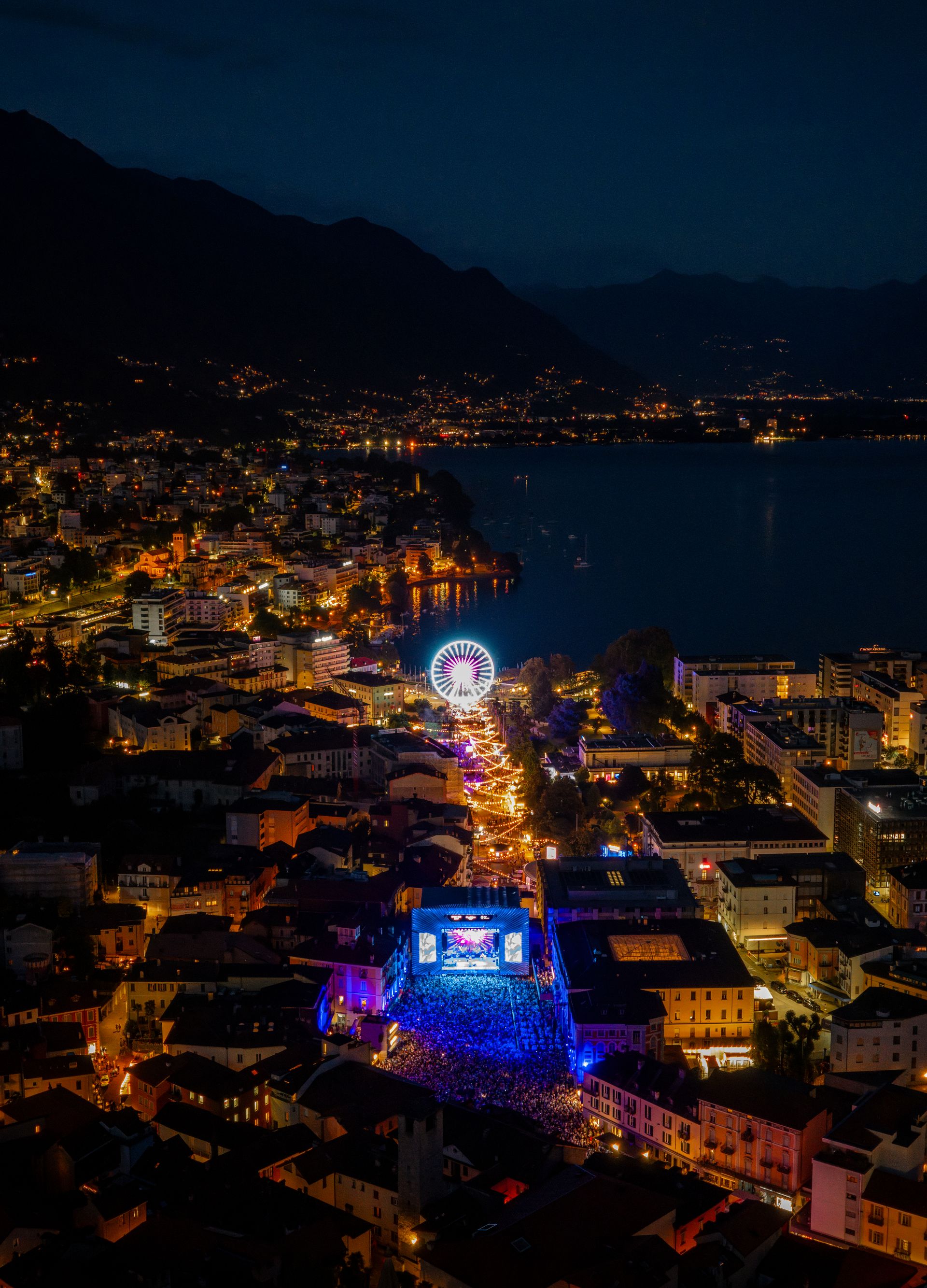 Veduta aerea notturna di Locarno con l’area illuminata del festival Moon+Stars, la ruota panoramica e il Lago Maggiore