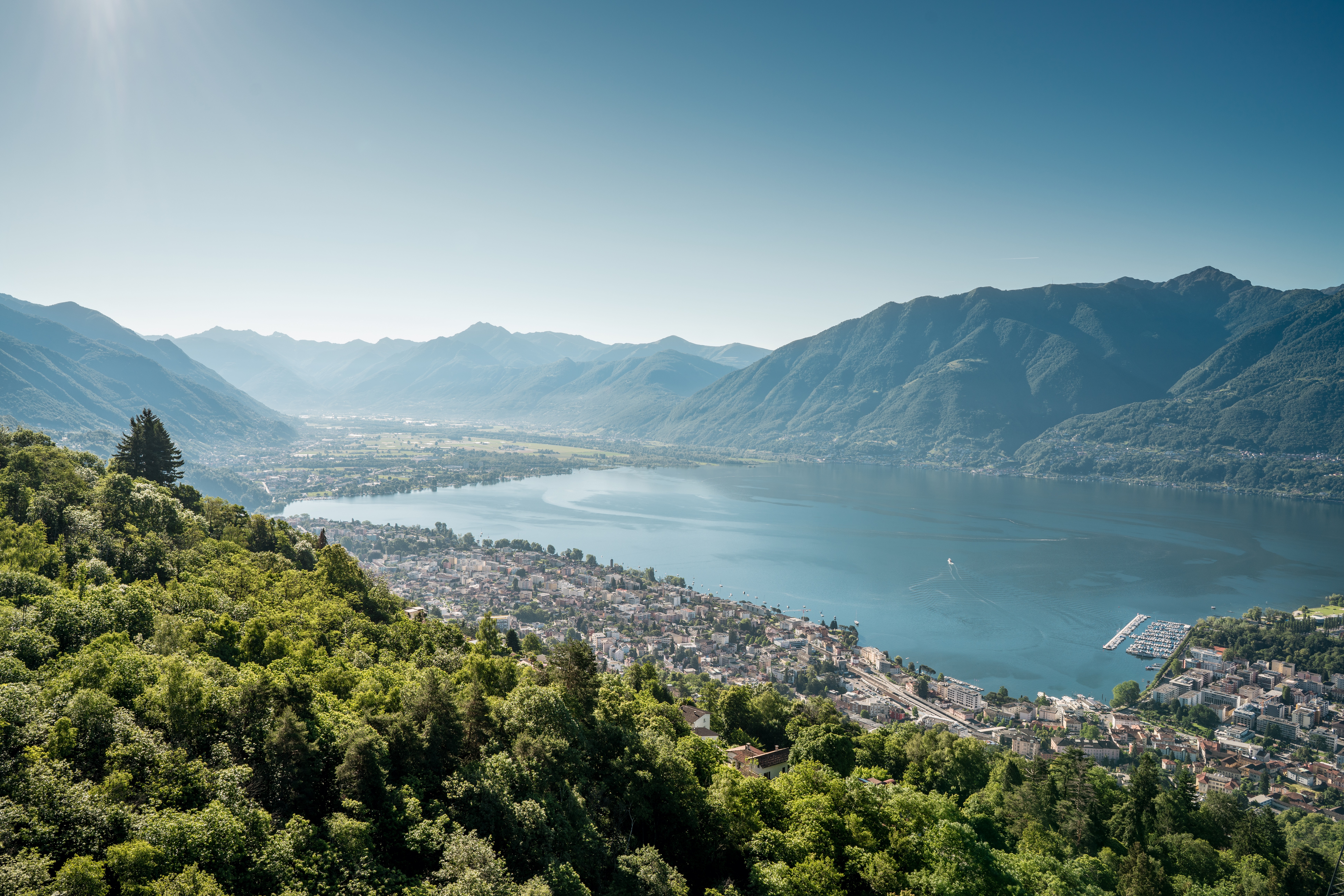 Panoramablick von Cardada auf Locarno und den Lago Maggiore an einem sonnigen Tag. Die Stadt liegt eingebettet zwischen grünen Wäldern und den Bergen des Tessins.