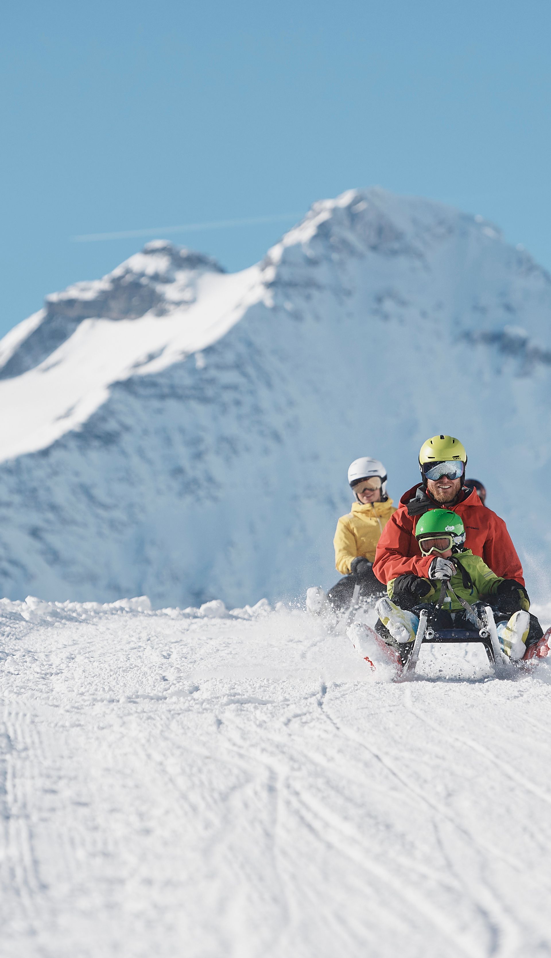 Group sledding on a snow-covered trail in Elm with alpine mountain scenery in the background.