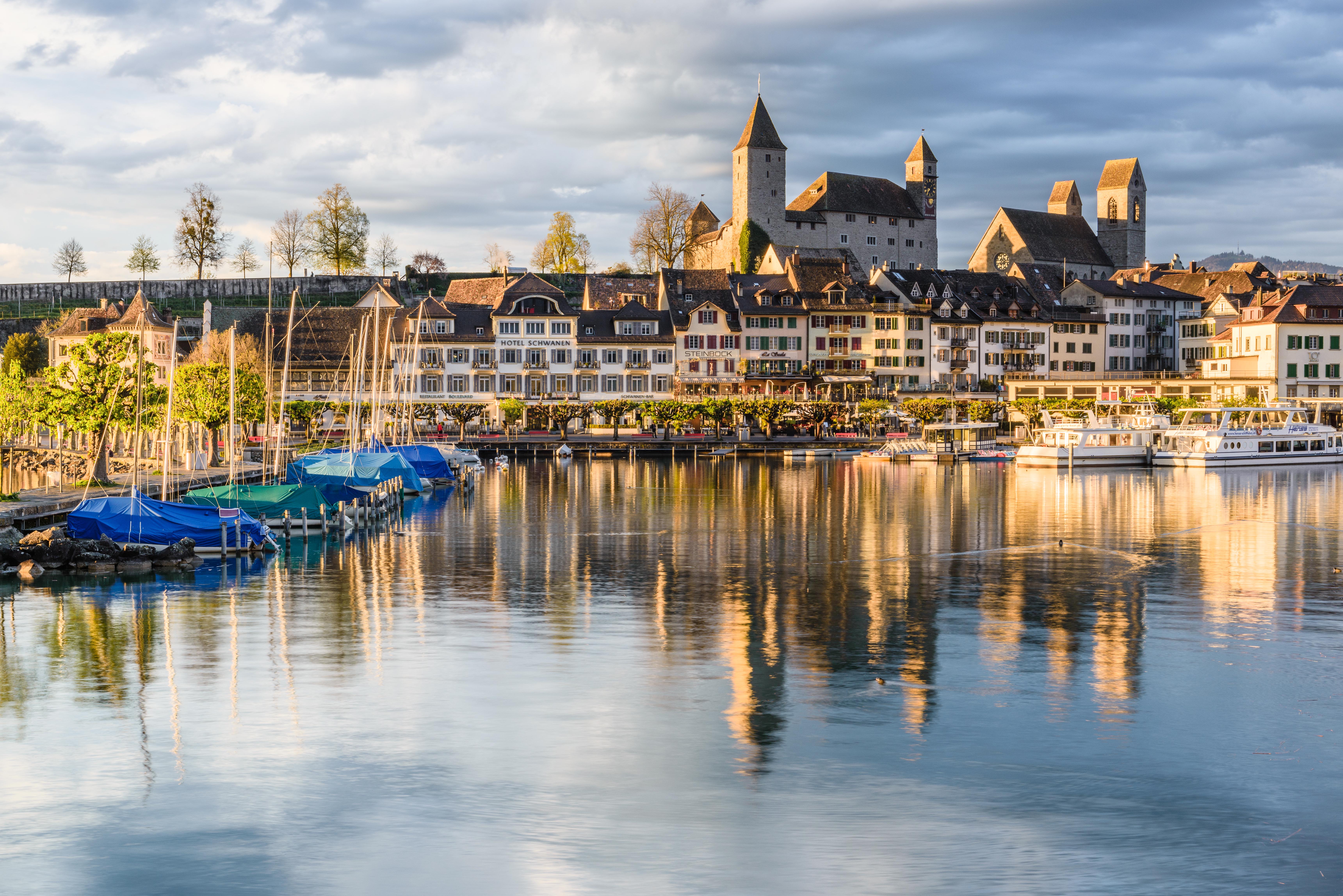 Vue sur le port de Rapperswil avec ses bateaux, ses maisons historiques et le château de Rapperswil baigné de lumière dorée.