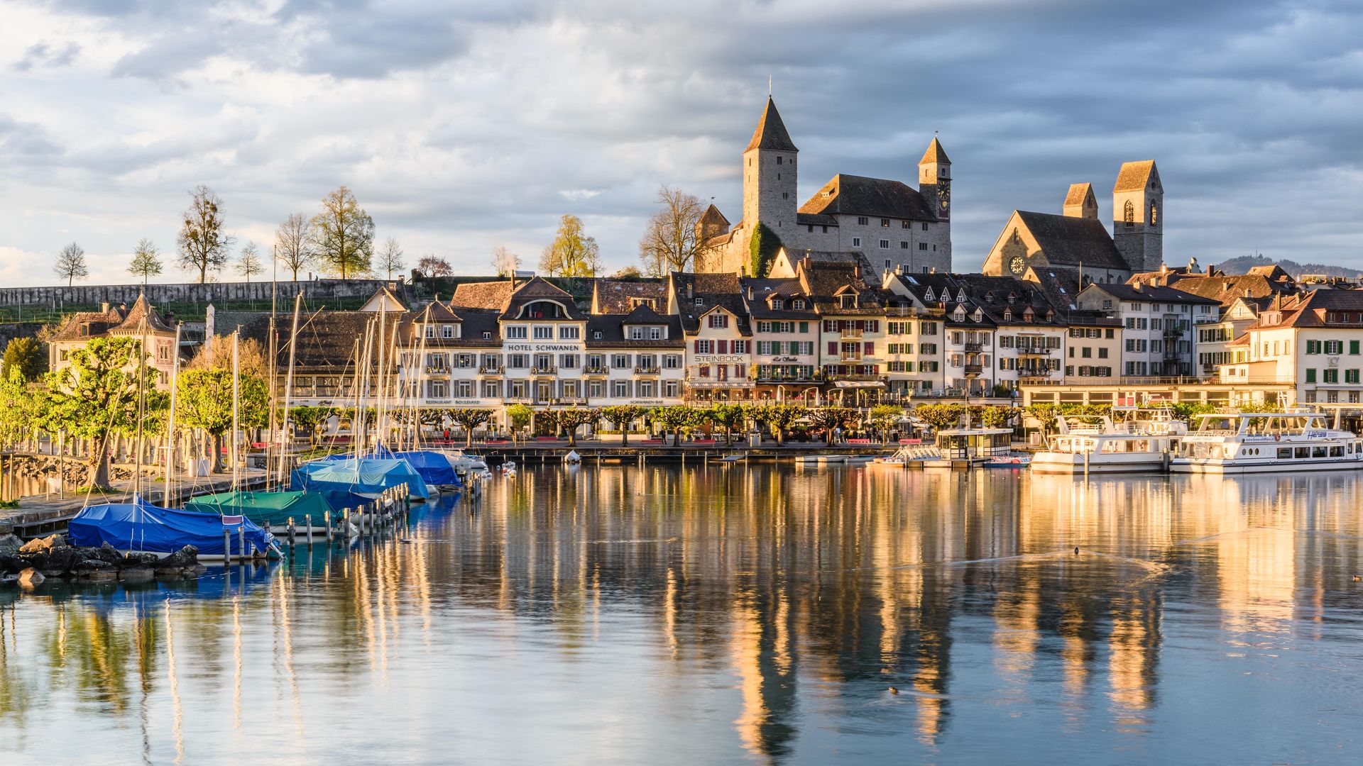 Vista sul porto di Rapperswil con barche, case storiche e il castello di Rapperswil illuminato dalla luce serale.