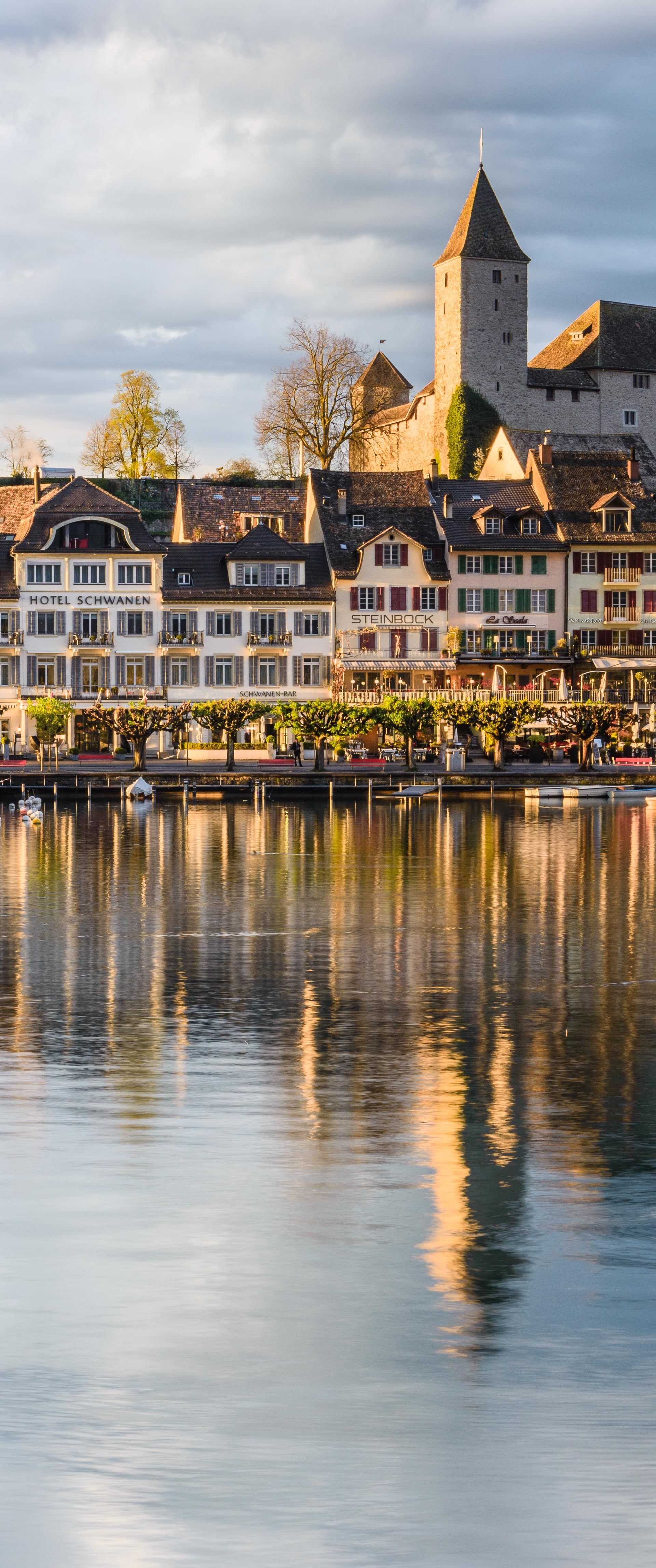 Blick auf den Hafen von Rapperswil mit Booten, historischen Häusern und dem Schloss Rapperswil im warmen Abendlicht.