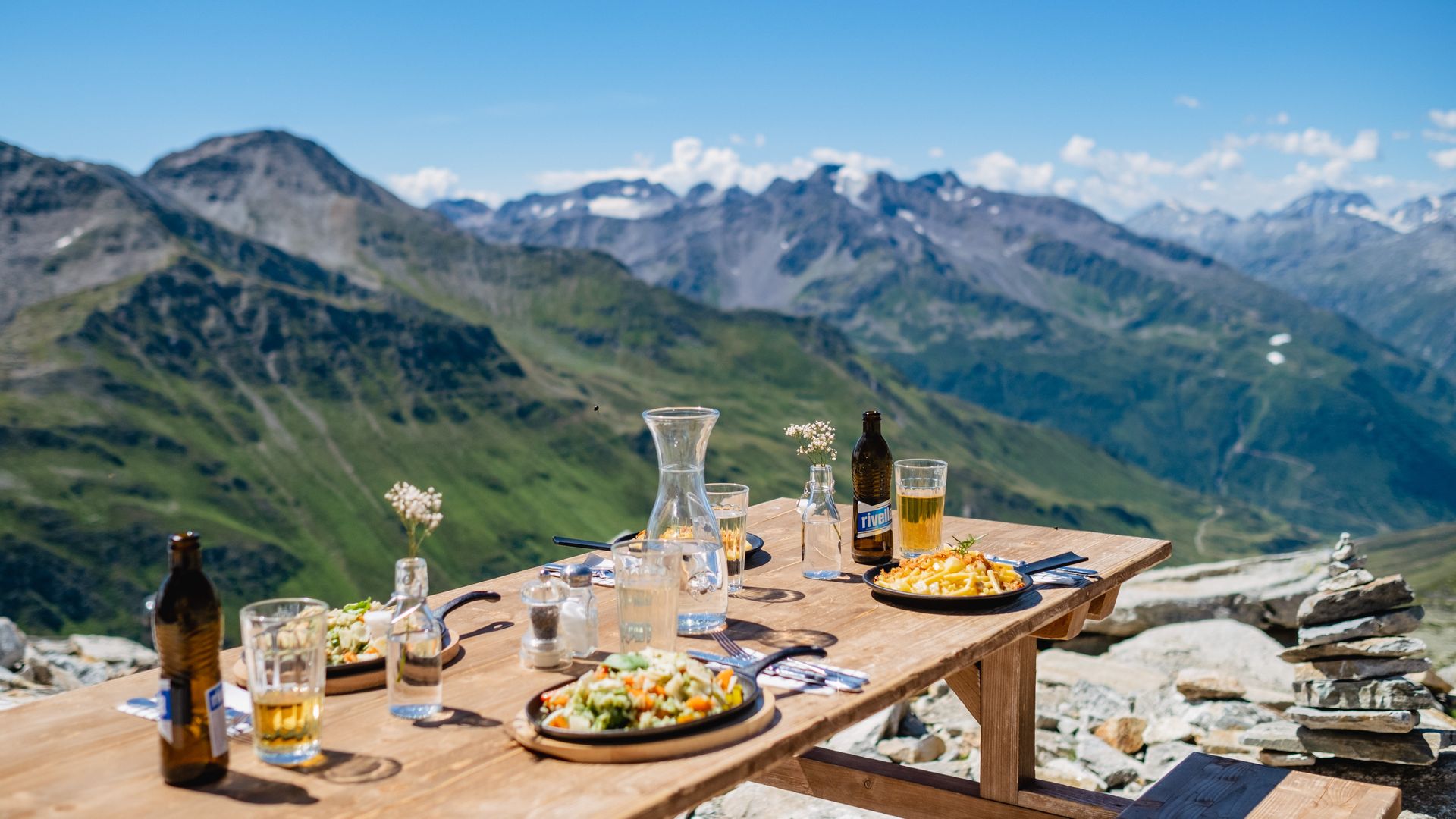 Wooden table with regional dishes and drinks on the terrace of the Schneehüenerstock restaurant above Andermatt, offering a breathtaking view of the surrounding Alps.