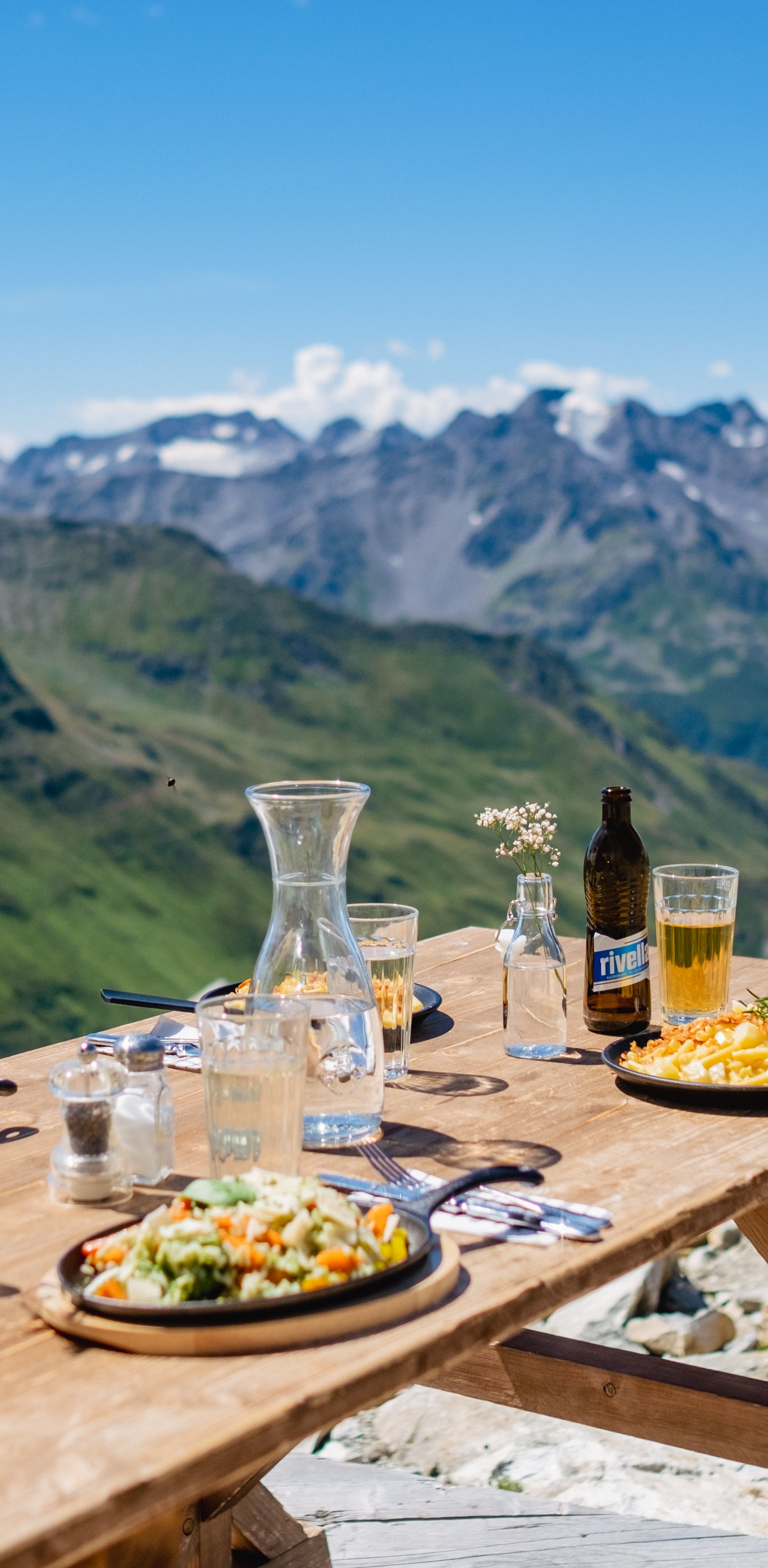 Wooden table with regional dishes and drinks on the terrace of the Schneehüenerstock restaurant above Andermatt, offering a breathtaking view of the surrounding Alps.