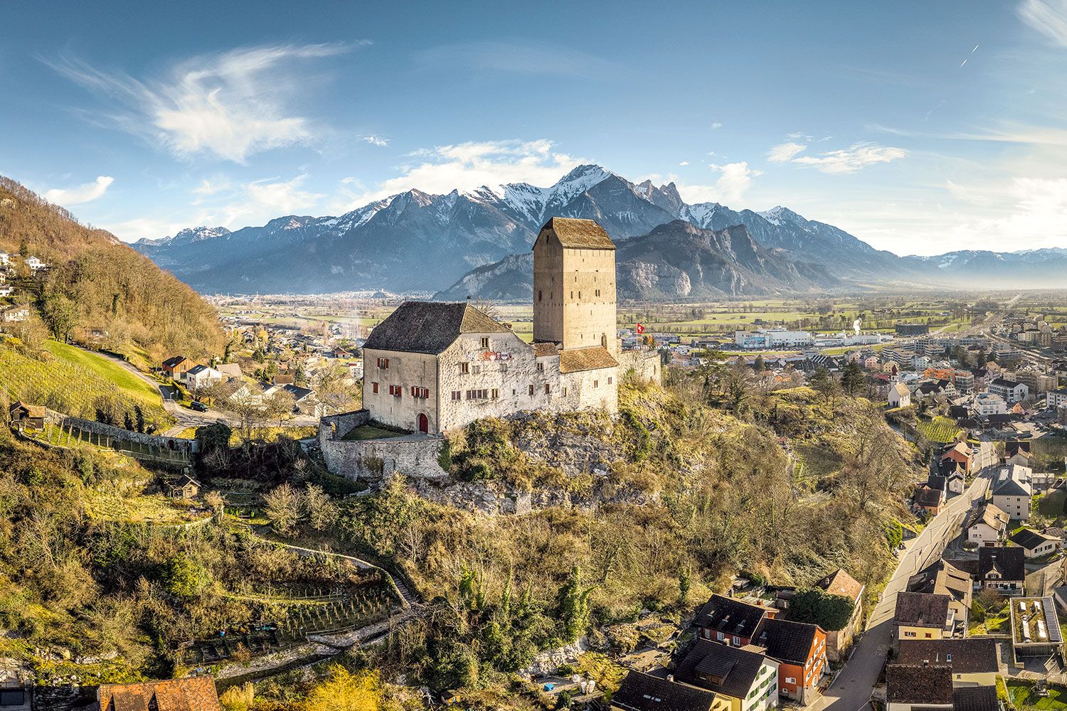 Sargans Castle perched on a rocky hill overlooking the town and the Rhine Valley, with snow-covered mountains in the background.