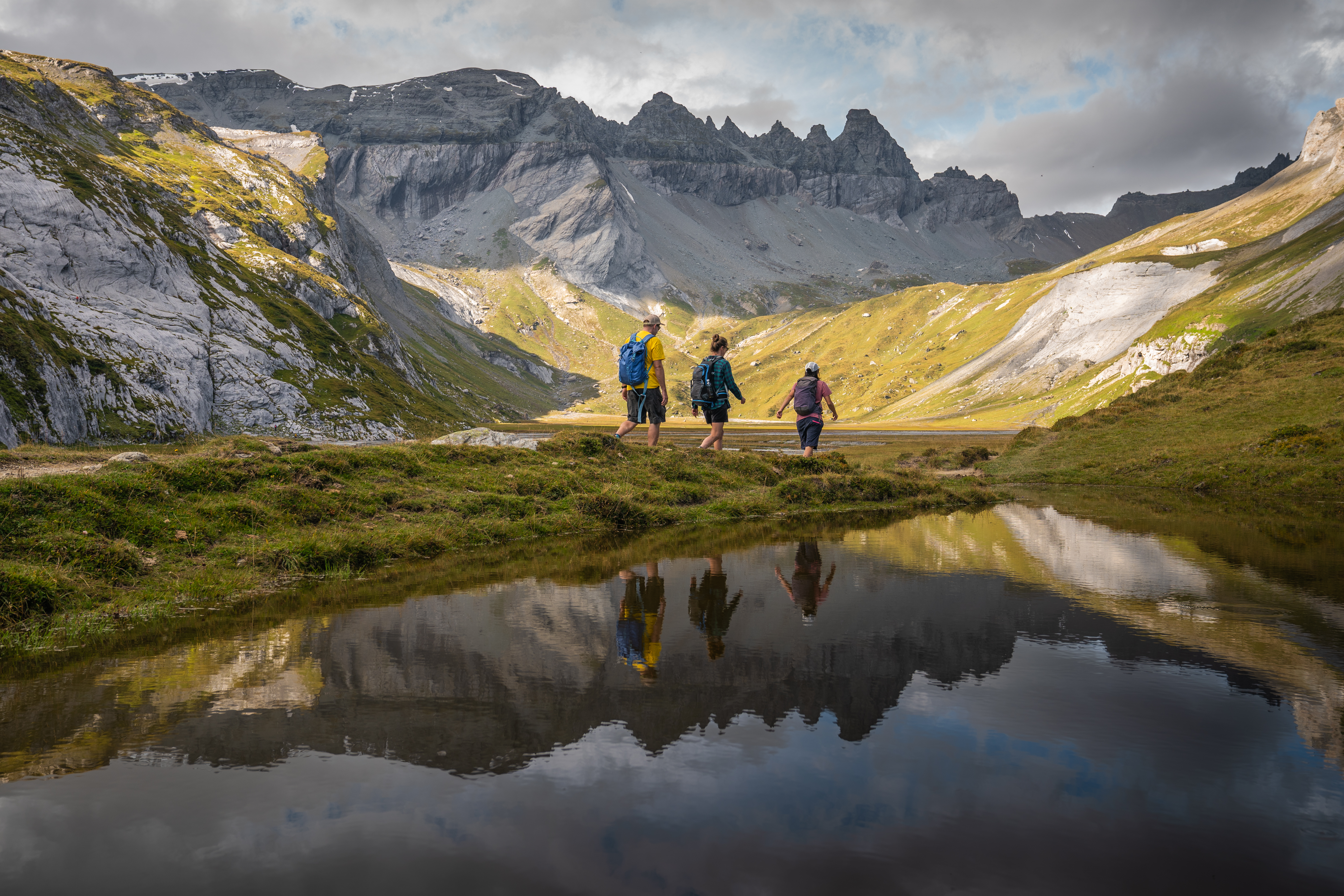 Wandernde auf der Segnesebene oberhalb von Flims in der Tektonikarena Sardona, umgeben von grauen Felswänden und grünen Alpwiesen, mit Spiegelung der Berge im Wasser – eindrucksvolle Landschaft in Graubünden.