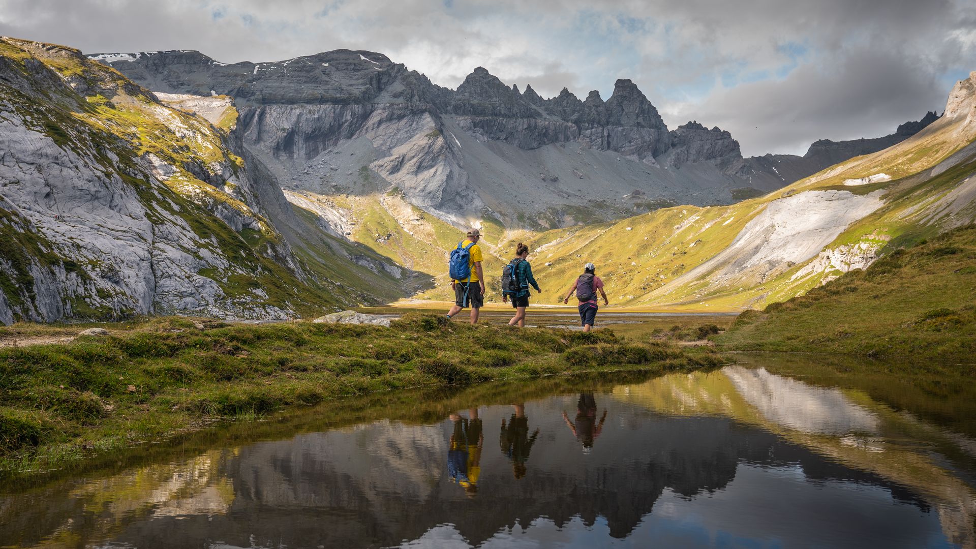 Wandernde auf der Segnesebene oberhalb von Flims in der Tektonikarena Sardona, umgeben von grauen Felswänden und grünen Alpwiesen, mit Spiegelung der Berge im Wasser – eindrucksvolle Landschaft in Graubünden.