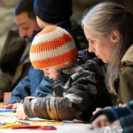 Un enfant avec une casquette orange et blanche se concentre sur son bricolage à côté d'un parent assis à une table.
