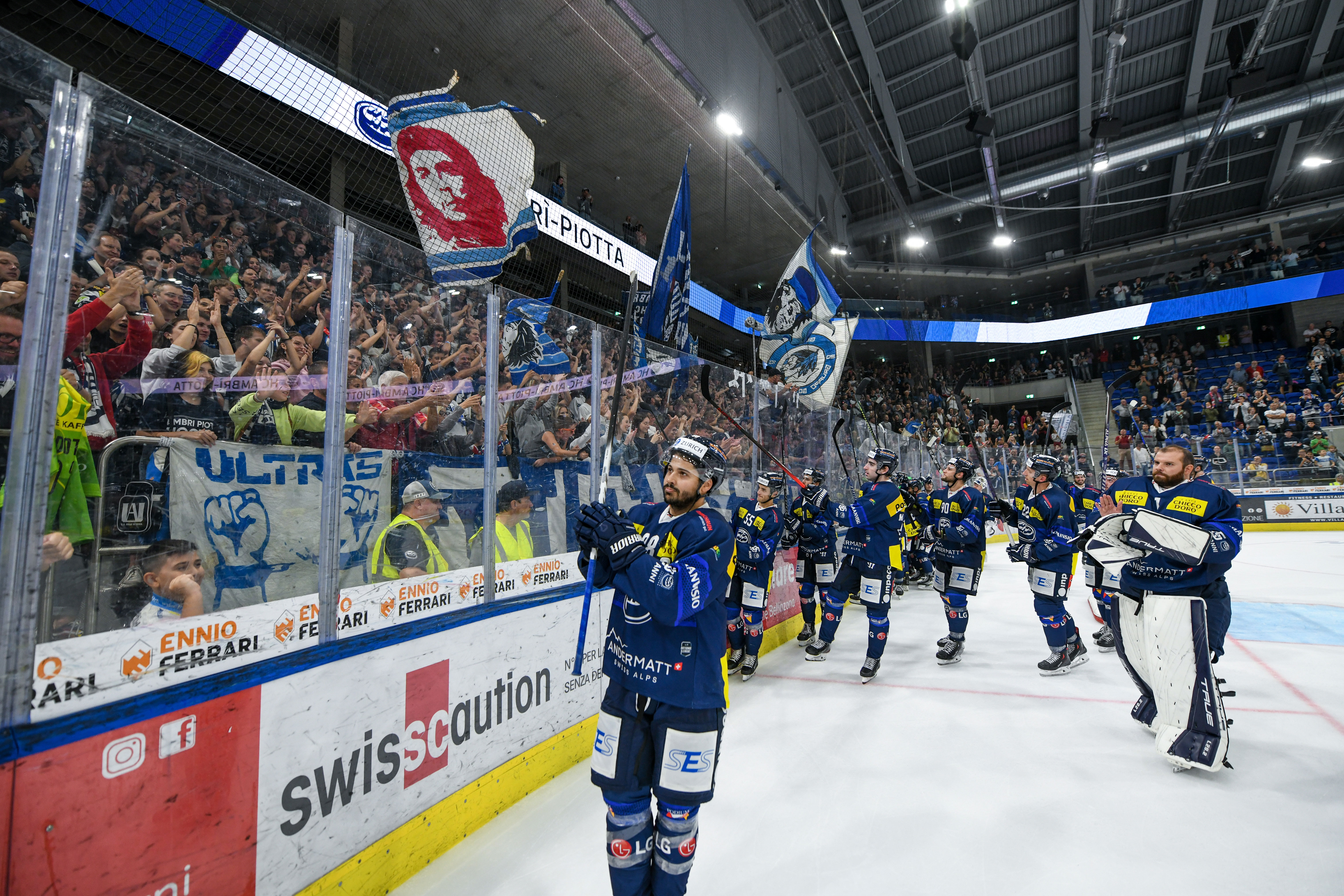 Die Spieler des HC Ambrì-Piotta bedanken sich nach dem Spiel bei ihren Fans in der Gottardo Arena. Im Hintergrund jubeln die Anhänger mit Fahnen und Bannern in Blau und Weiss.
