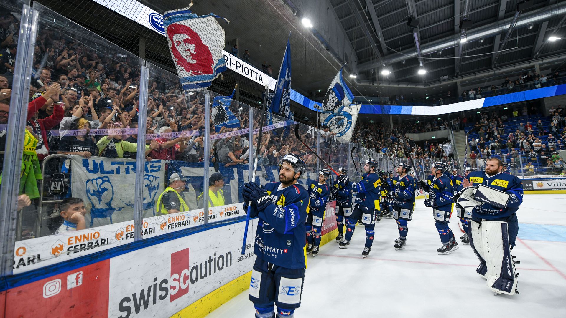 I giocatori dell’HC Ambrì-Piotta ringraziano i tifosi dopo la partita nella Gottardo Arena, mentre sugli spalti sventolano bandiere e striscioni biancoblù.