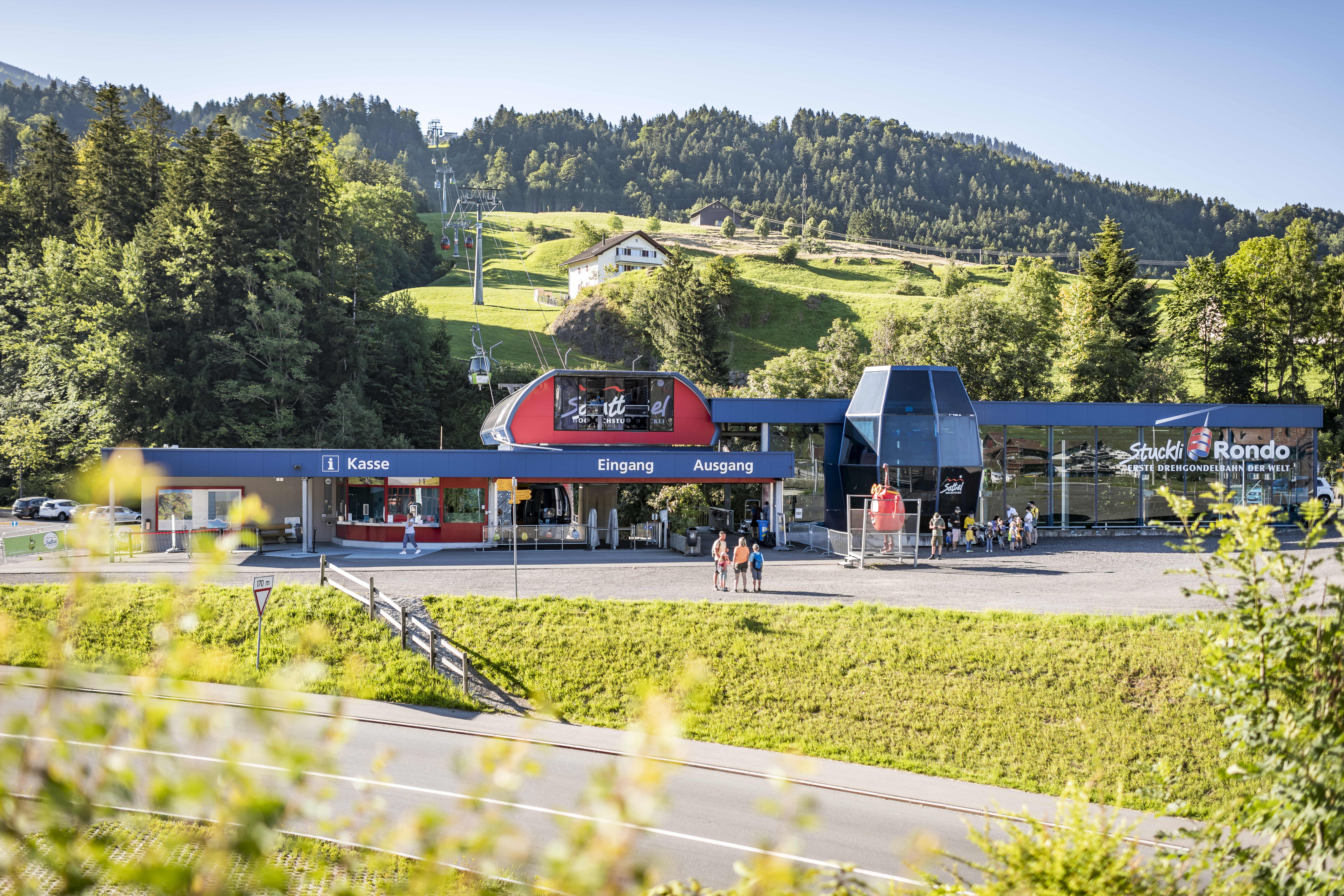 Frontansicht der Bergbahnstation mit wartenden Personen vor dem Einstieg in die Gondelbahn.