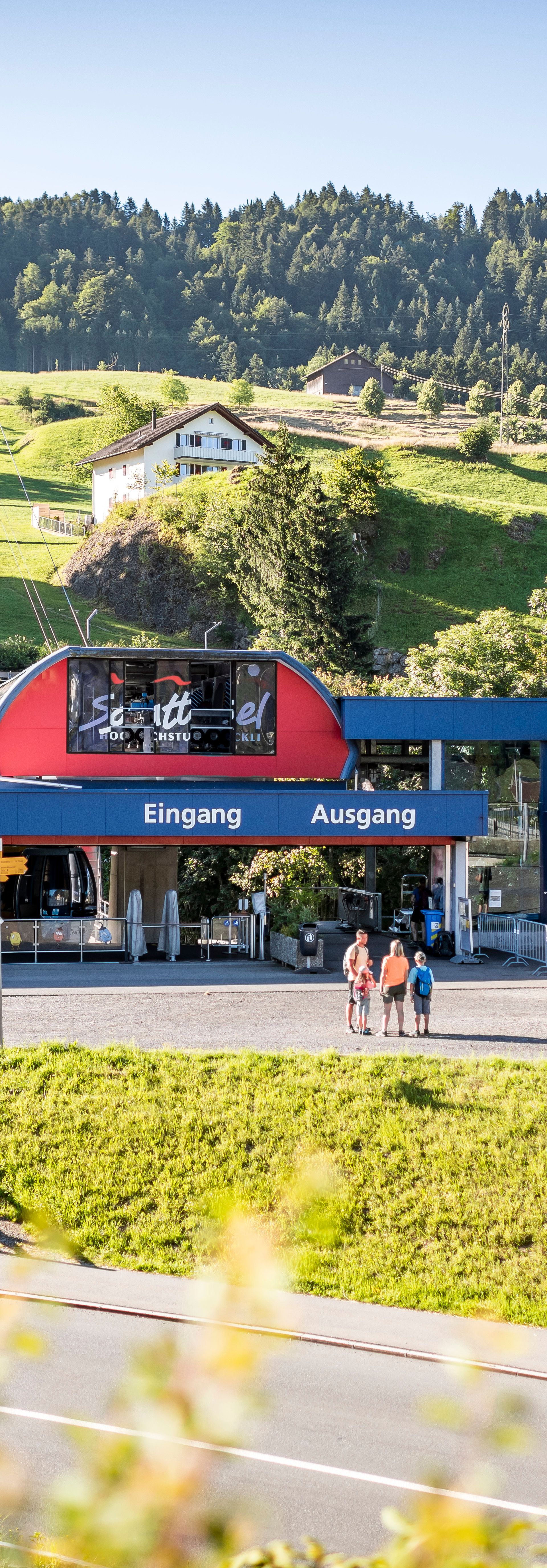 Frontansicht der Bergbahnstation mit wartenden Personen vor dem Einstieg in die Gondelbahn.