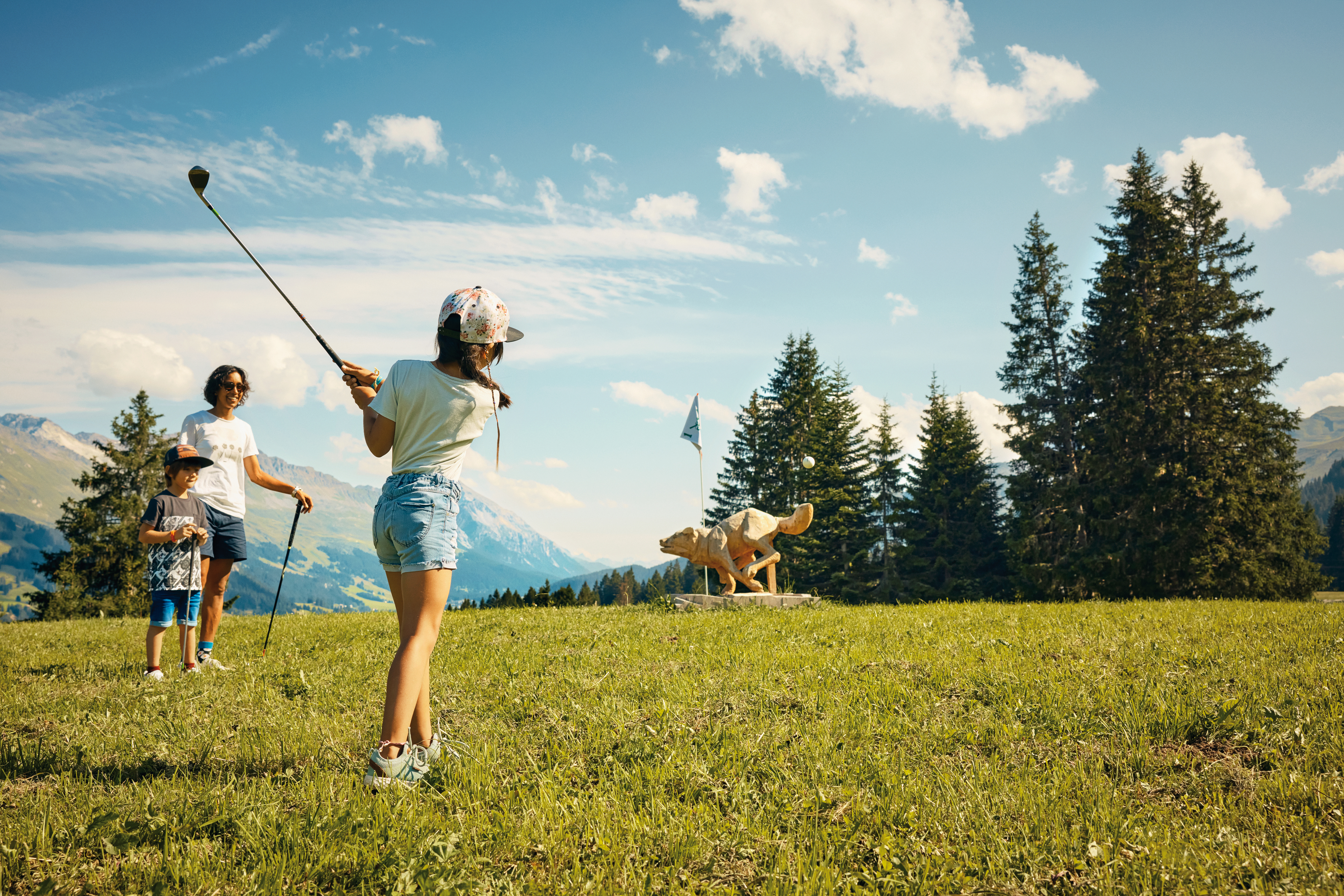 Ragazza che gioca a cross golf con la famiglia a Brambrüesch vicino a Coira, con panorama alpino e scultura in legno