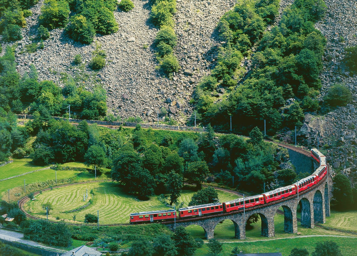 Roter Zug fährt um Kurve vor einem Hang mit Geröll und grünen Bäumen