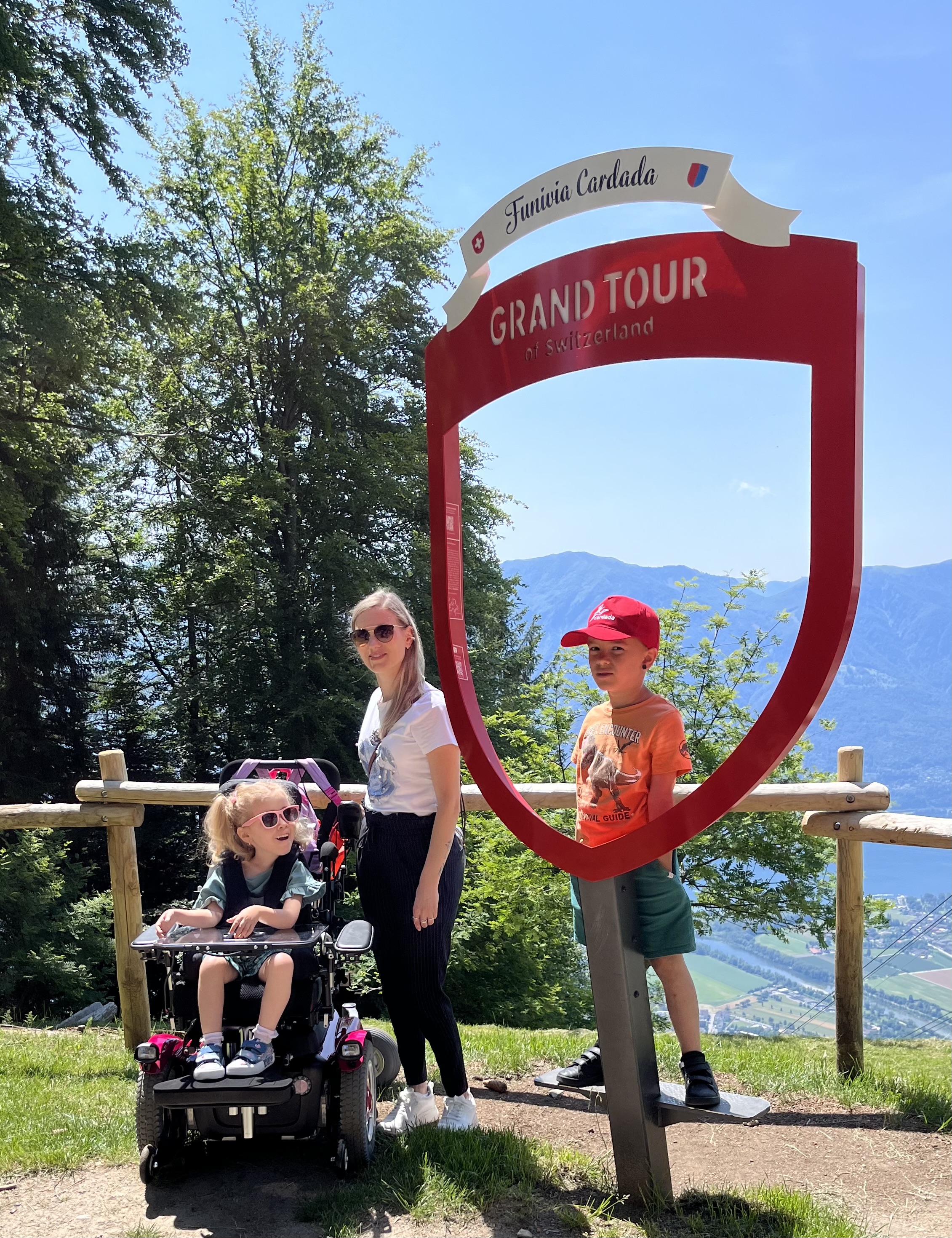 Eine Familie posiert beim Grand Tour of Switzerland-Rahmen auf Cardada oberhalb von Locarno. Das Kind im Rollstuhl lächelt, im Hintergrund die Magadinoebene und die Tessiner Berge.