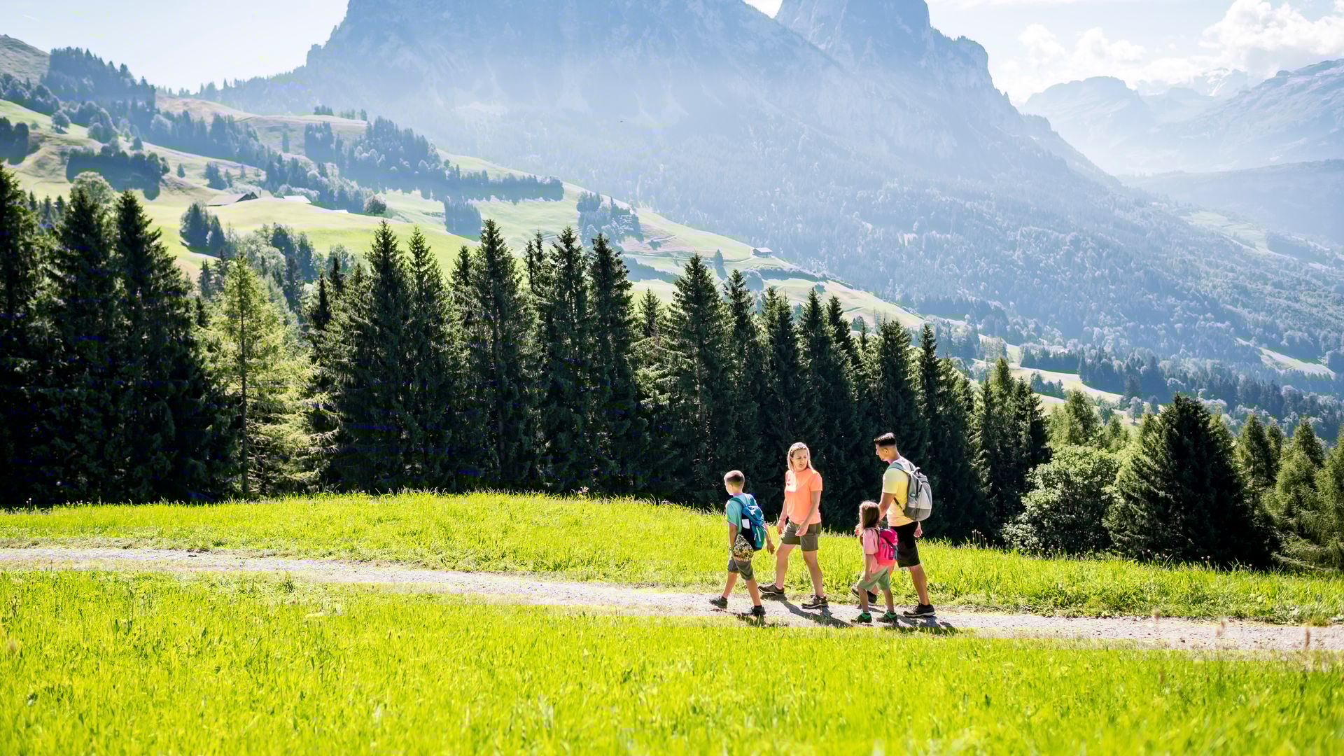 Familie wandert auf einem sonnigen Weg bei Engelstock Sattel-Hochstuckli mit Blick auf grüne Wälder, Wiesen und die Schwyzer Alpen.