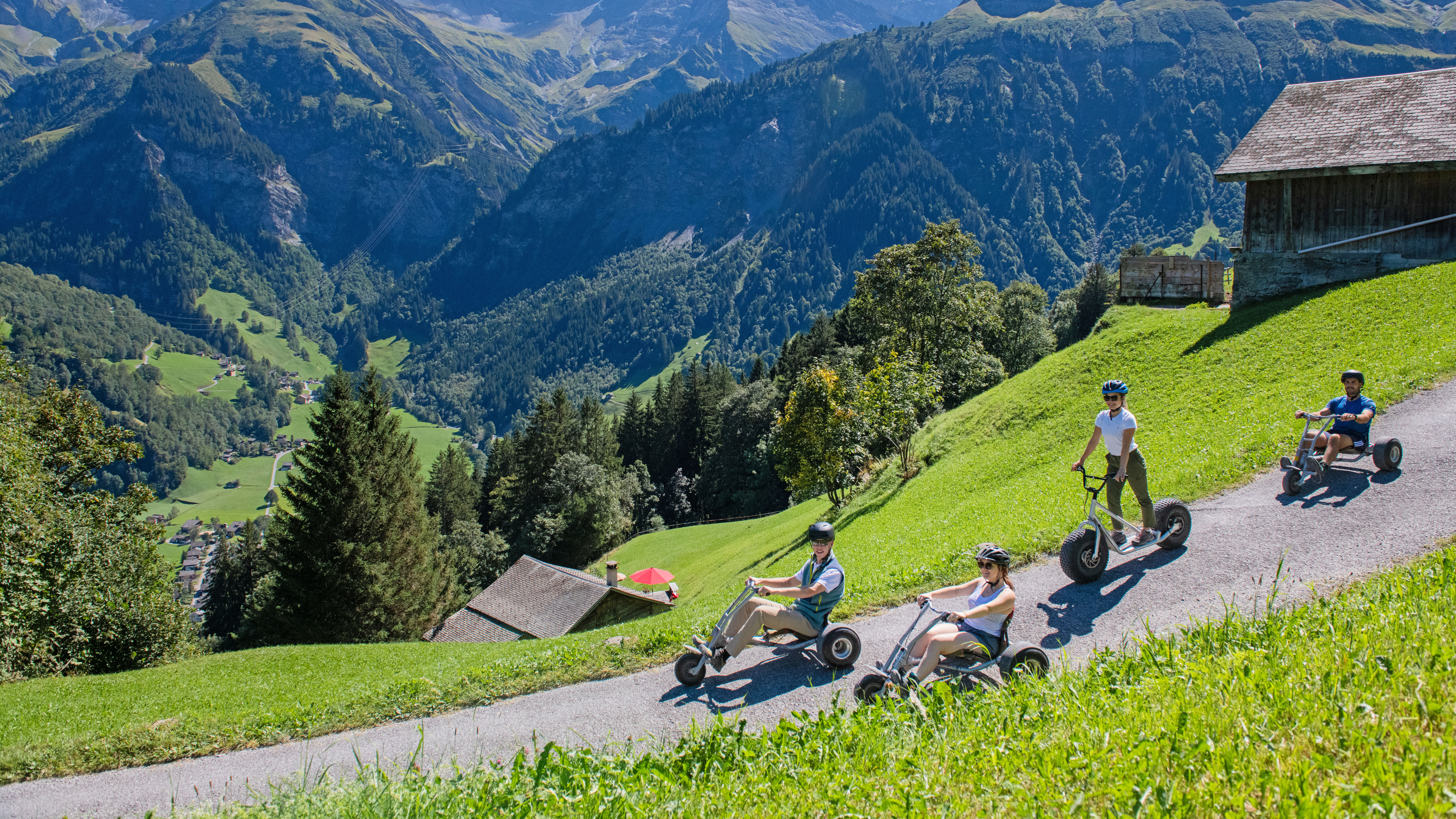Ausflügler fahren mit Trottinetts und Mountaincarts auf der Panoramastrasse bei den Sportbahnen Elm, umgeben von den imposanten Glarner Alpen an einem sonnigen Tag