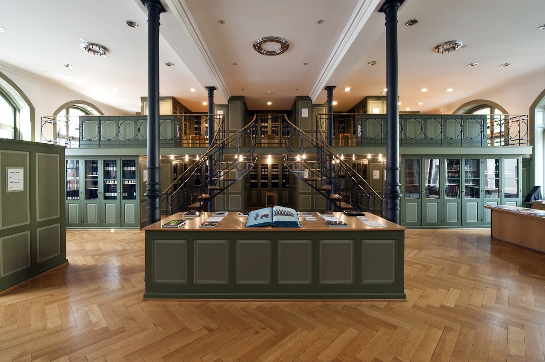 Vue intérieure de la bibliothèque historique du Musée du textile de Saint-Gall, avec table d’exposition centrale, escalier en colimaçon et rayonnages verts sur deux niveaux.