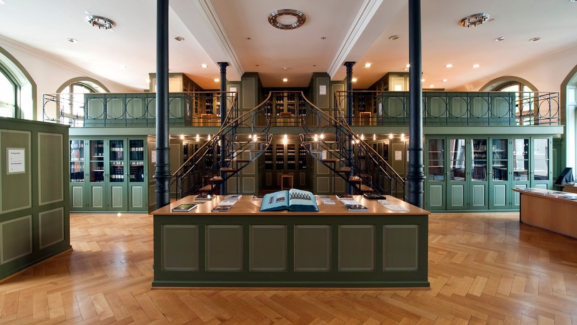 Innenansicht der historischen Bibliothek im Textilmuseum St. Gallen mit zentralem Ausstellungstisch, Wendeltreppe und grüner Regalwand auf zwei Ebenen.