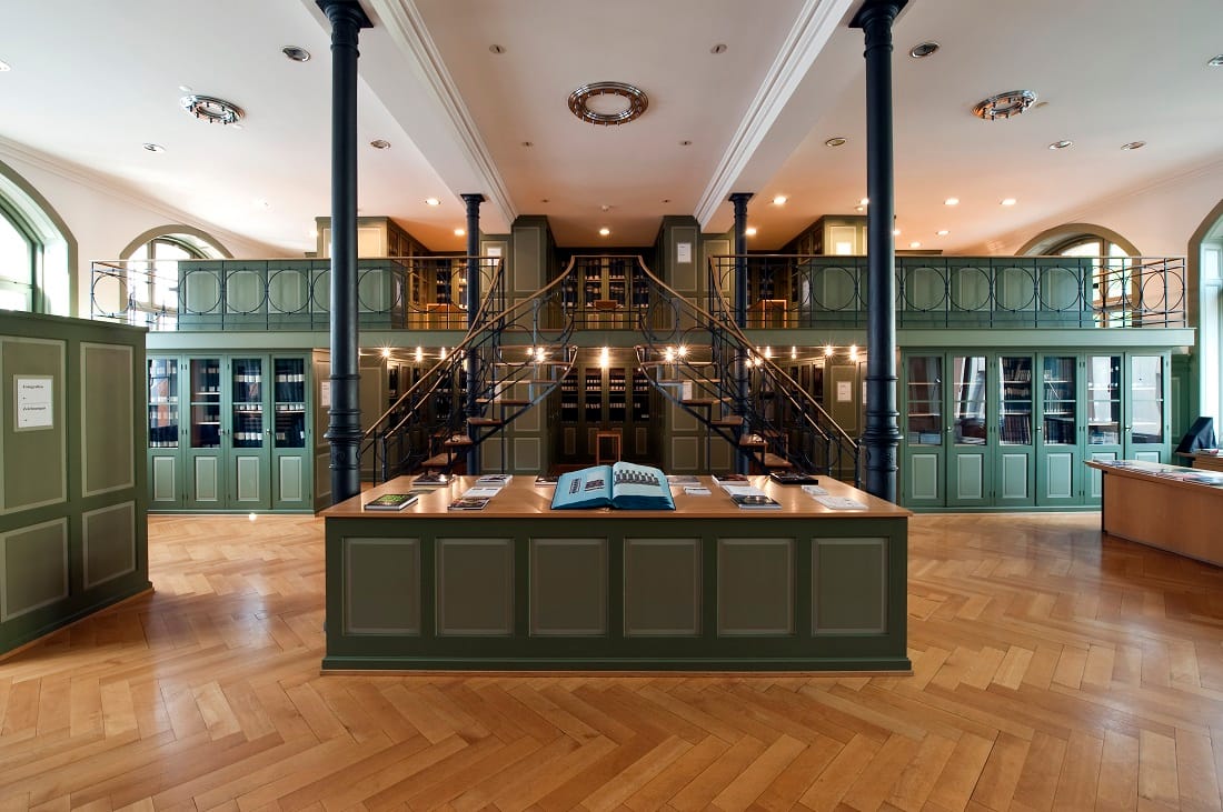 Vista interna della biblioteca storica del Museo del Tessile di San Gallo, con tavolo espositivo centrale, scala a chiocciola e scaffalature verdi su due livelli.