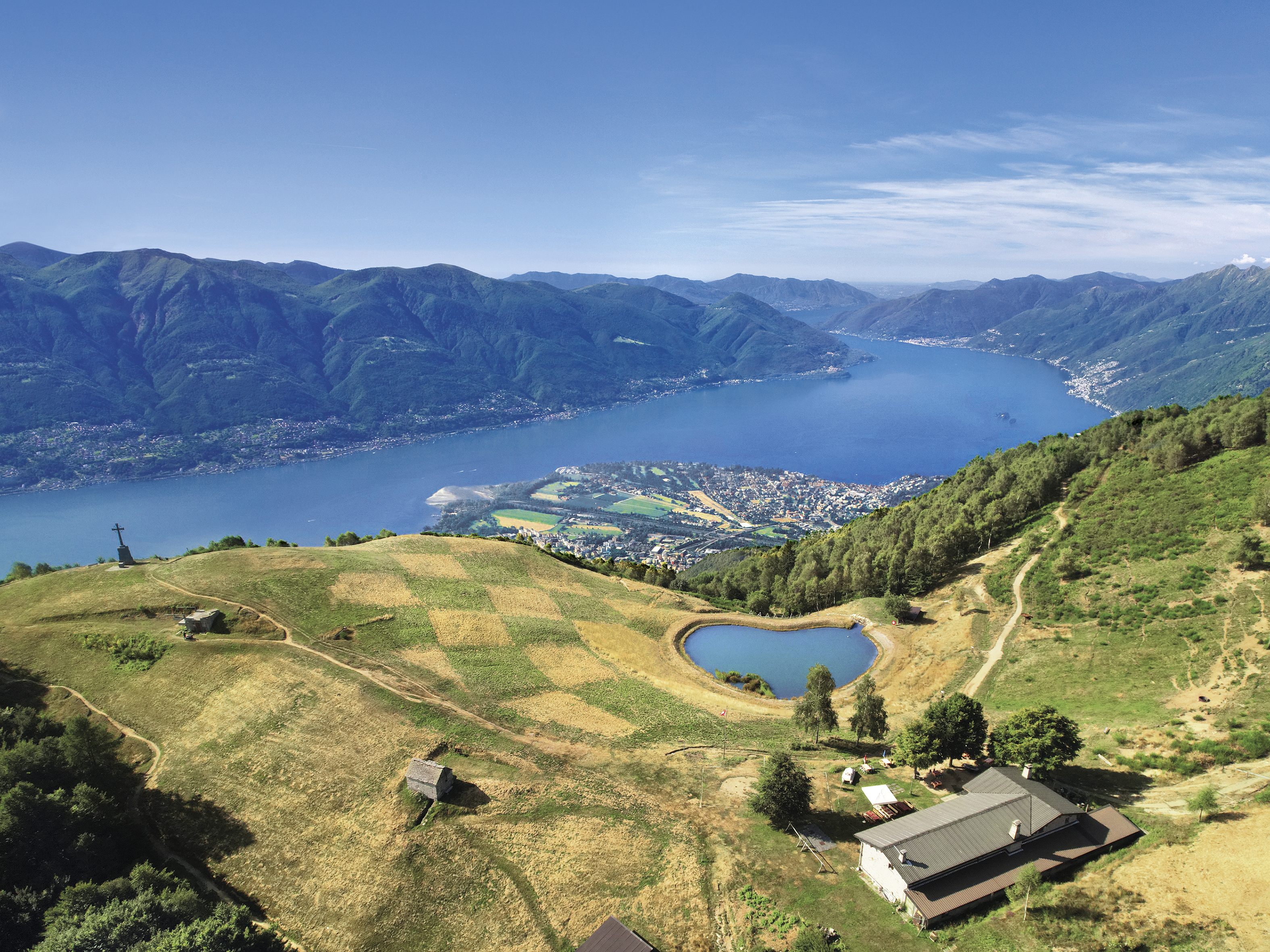 Sommerliches Panorama von Cardada Cimetta mit Blick auf den Lago Maggiore, das kleine Herz-See-Becken und die Schachbrettlandschaft oberhalb von Locarno im Tessin.