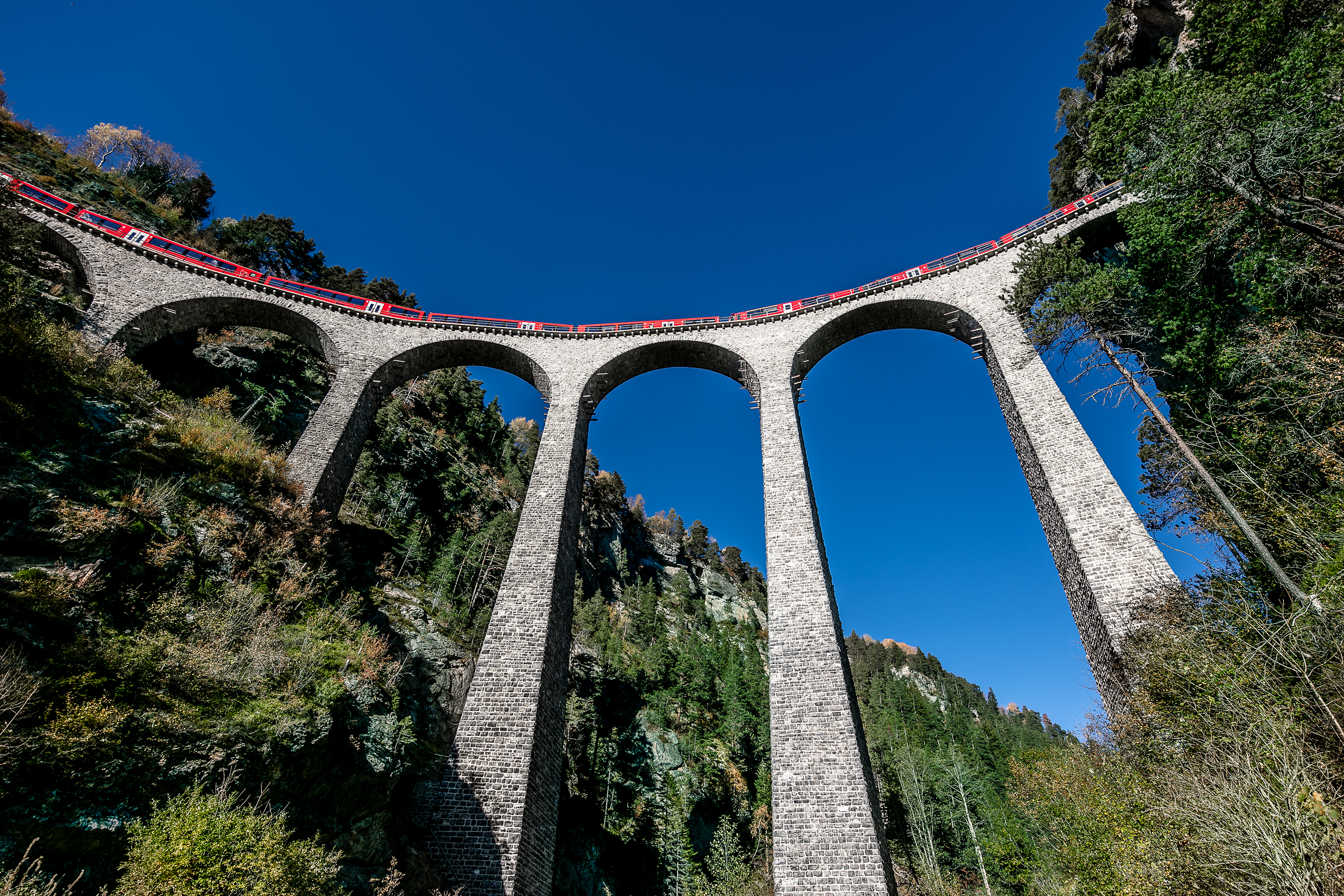 Un train rouge du Chemin de fer rhétique traverse l’impressionnant viaduc de Landwasser près de Filisur, dans le canton des Grisons, sous un ciel bleu profond.