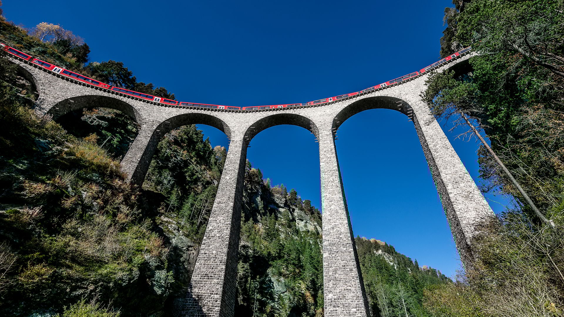 Un treno rosso della Ferrovia Retica attraversa l’imponente Viadotto Landwasser vicino a Filisur, nei Grigioni, sotto un cielo blu intenso.
