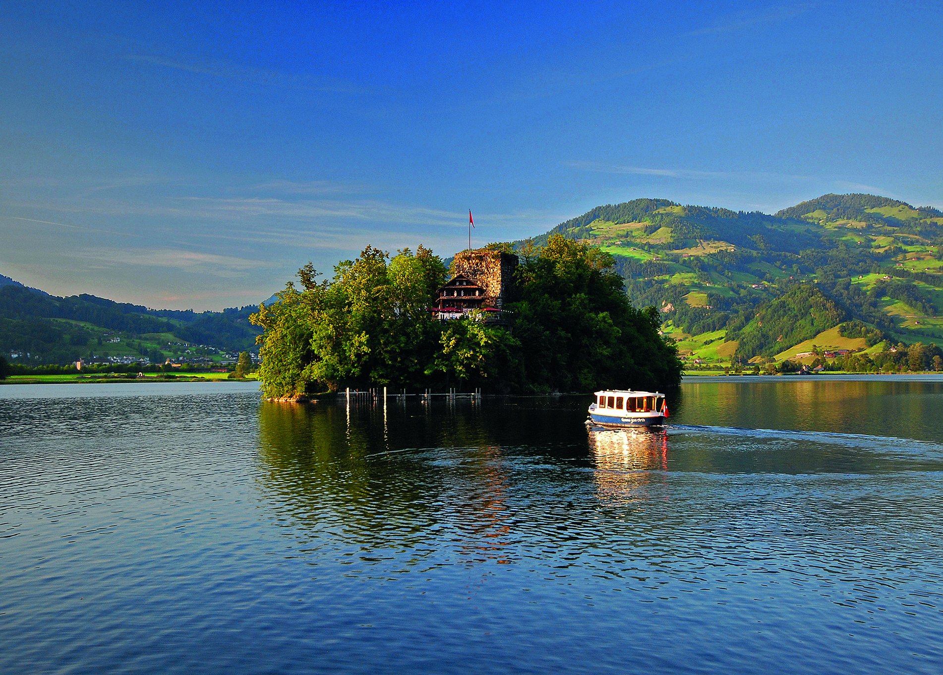 L’île de Schwanau sur le lac de Lauerz, avec son auberge historique, entourée de collines verdoyantes et d’un bateau sur le lac au soleil.