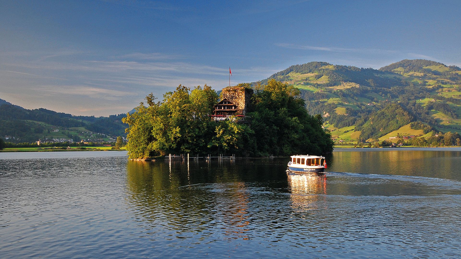 L’île de Schwanau sur le lac de Lauerz, avec son auberge historique, entourée de collines verdoyantes et d’un bateau sur le lac au soleil.