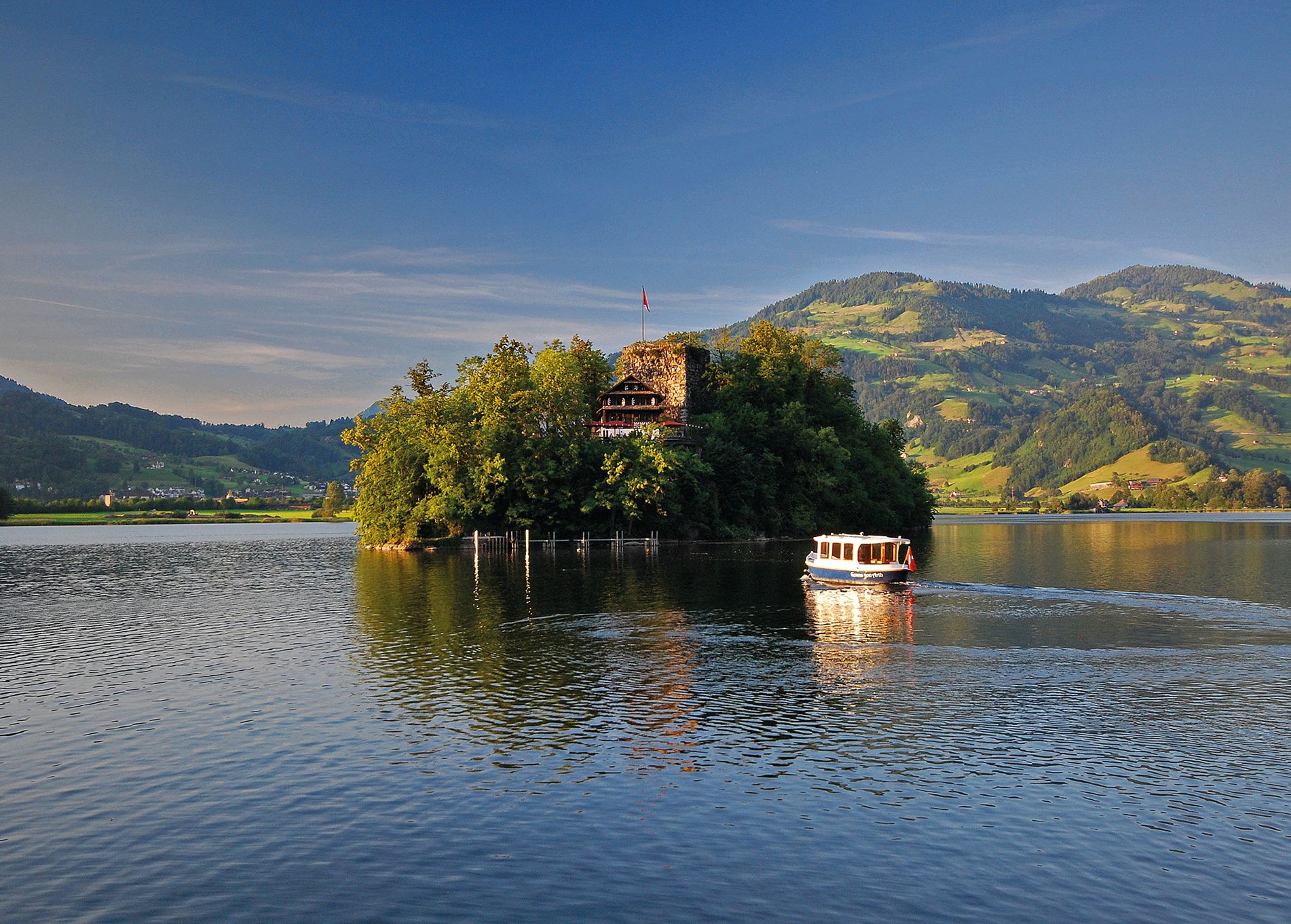 L’isola di Schwanau sul Lago di Lauerz con la sua locanda storica, circondata da colline verdi e una barca sul lago al sole.