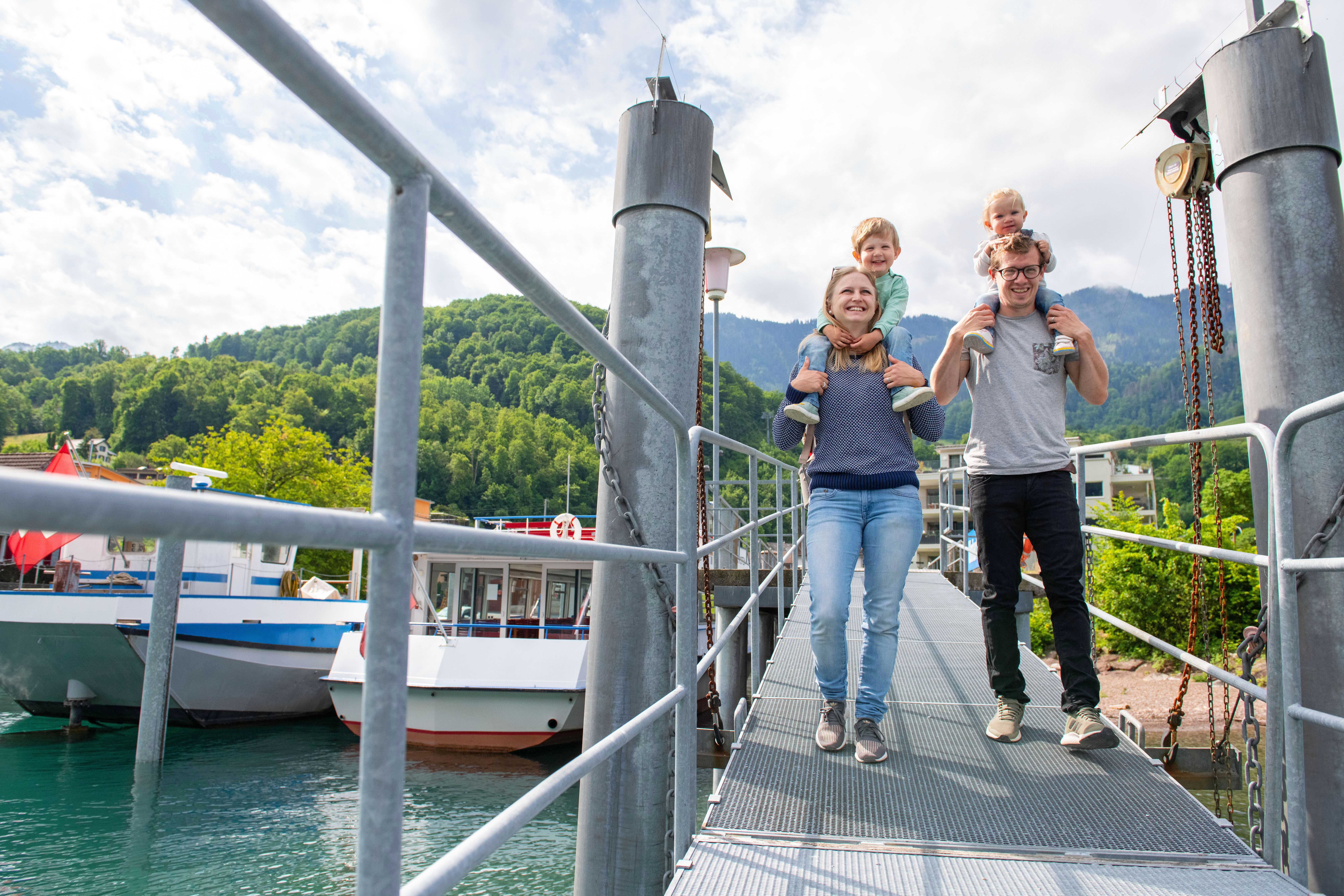 Familie mit 2 Kindern auf den Schultern läuft über einen Steg mit einem Schiff im Hintergrund