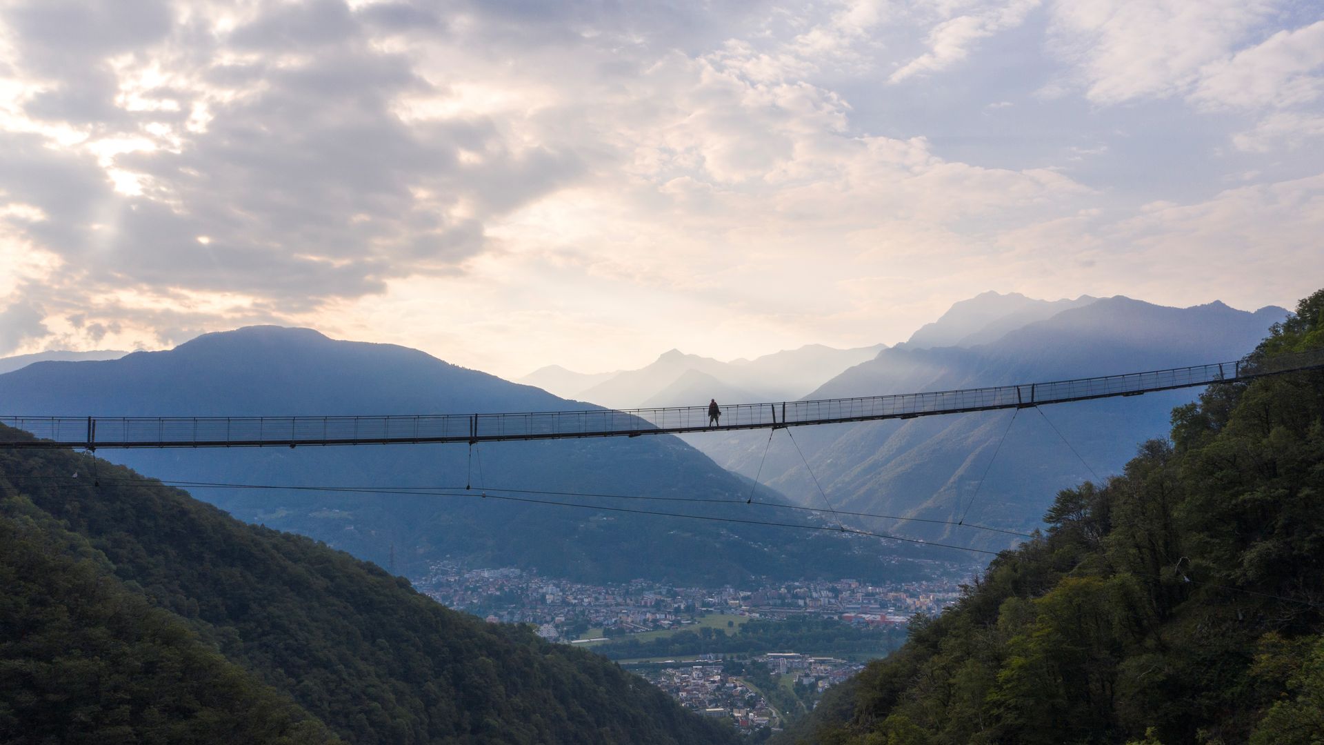 Person crossing the Ponte Tibetano Carasc suspension bridge near Monte Carasso with a panoramic view of the Ticino mountains and valley.