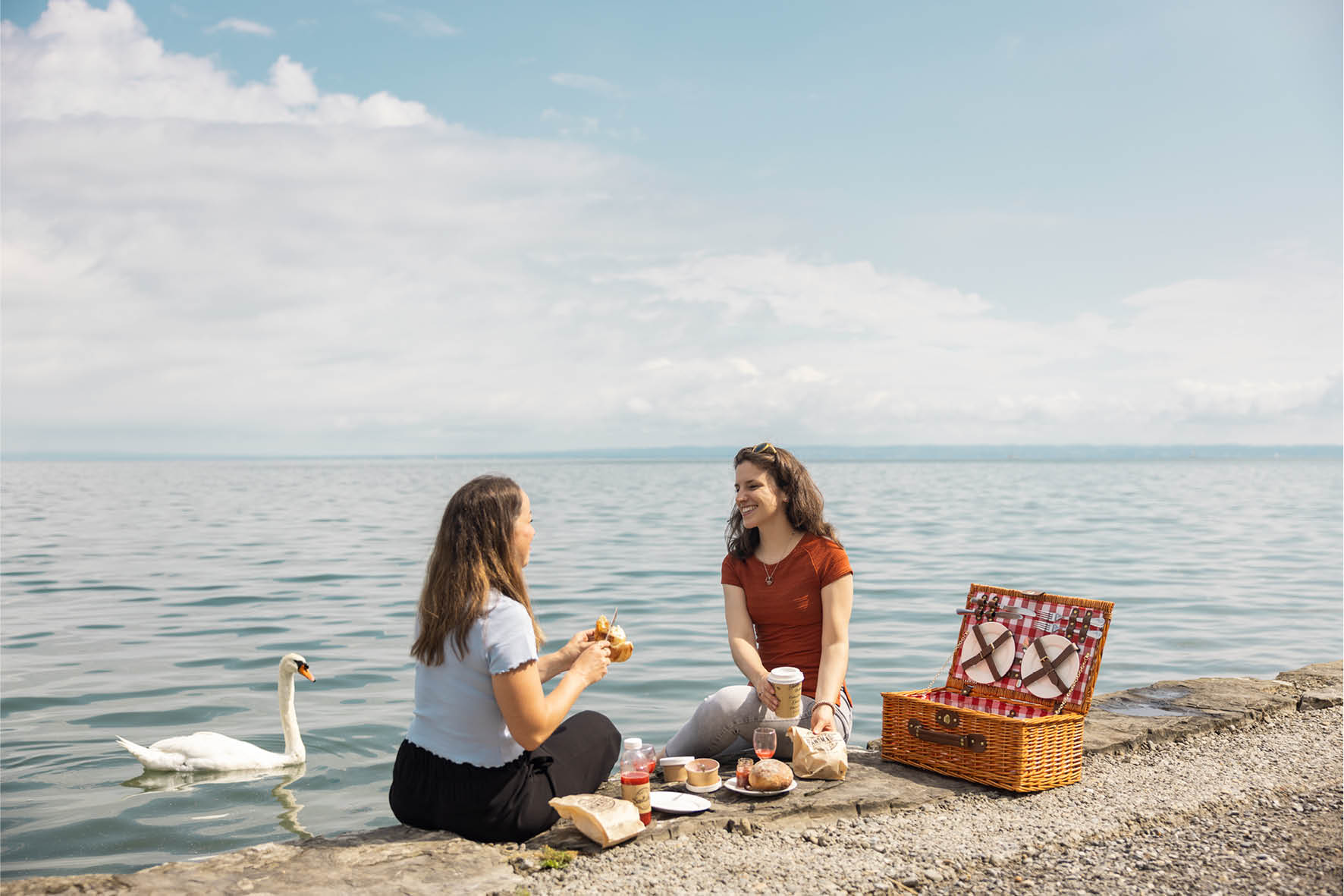 Zwei Frauen essen am Ufer des Bodensee's einen Brunch mit dem Zmorge-Korb. Auf dem See ist ein Schwan zu sehen. 