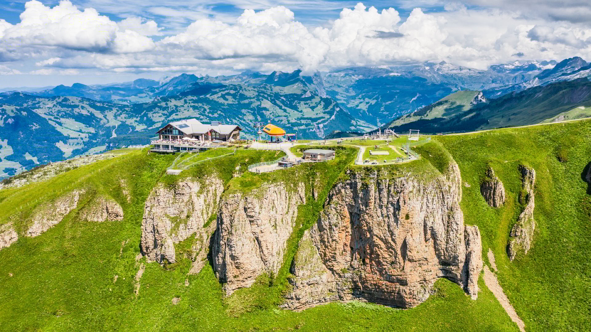 Vista sulla vetta del Fronalpstock sopra Stoos con il ristorante di montagna, la piattaforma panoramica e i paesaggi alpini verdi d’estate.