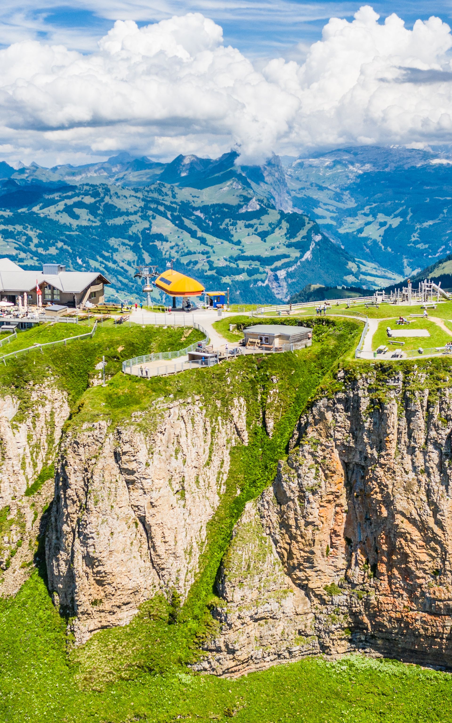 Vista sulla vetta del Fronalpstock sopra Stoos con il ristorante di montagna, la piattaforma panoramica e i paesaggi alpini verdi d’estate.