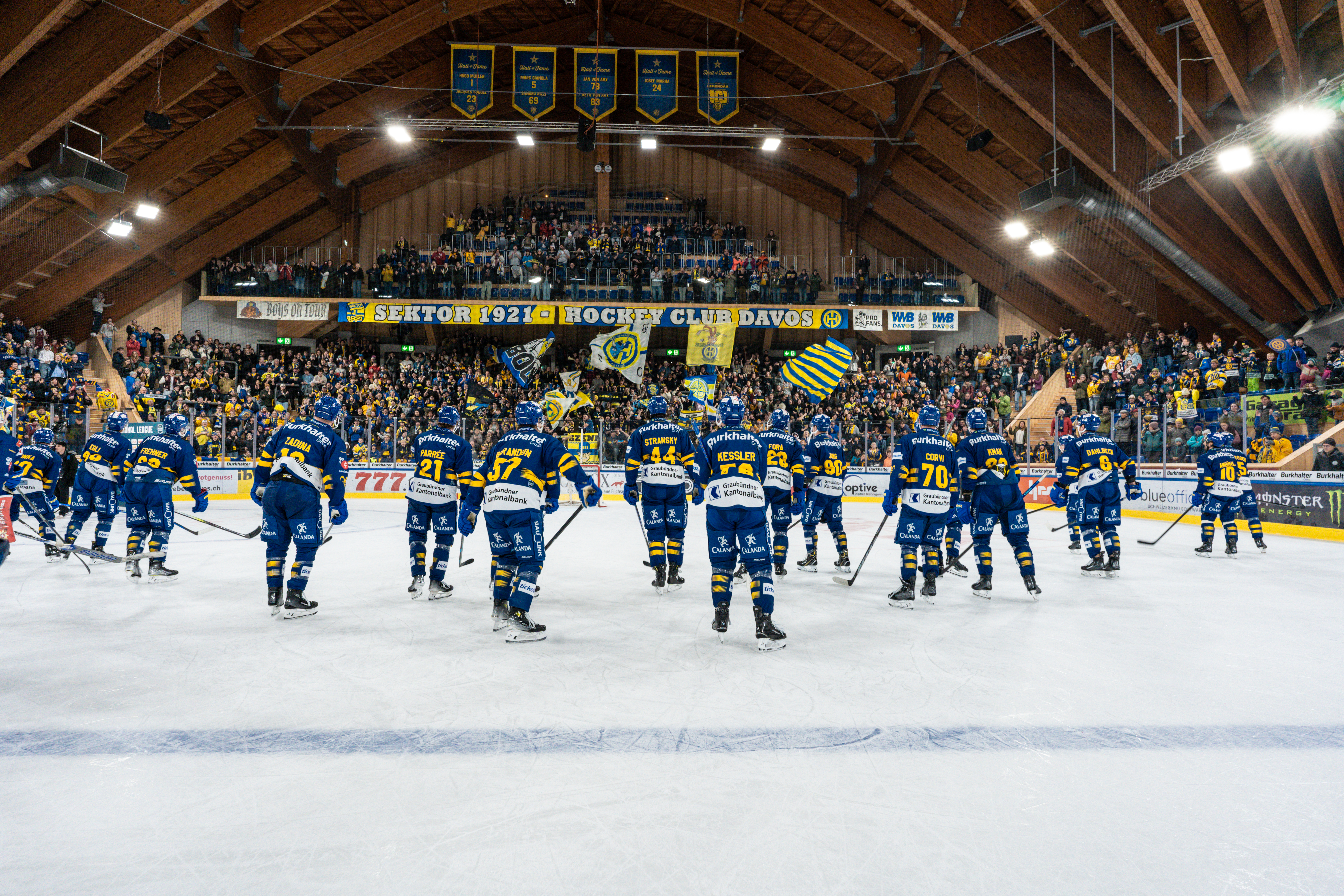 Players of HC Davos stand on the ice after the game, greeting their fans as the crowd waves yellow and blue flags in the iconic Davos Ice Stadium.