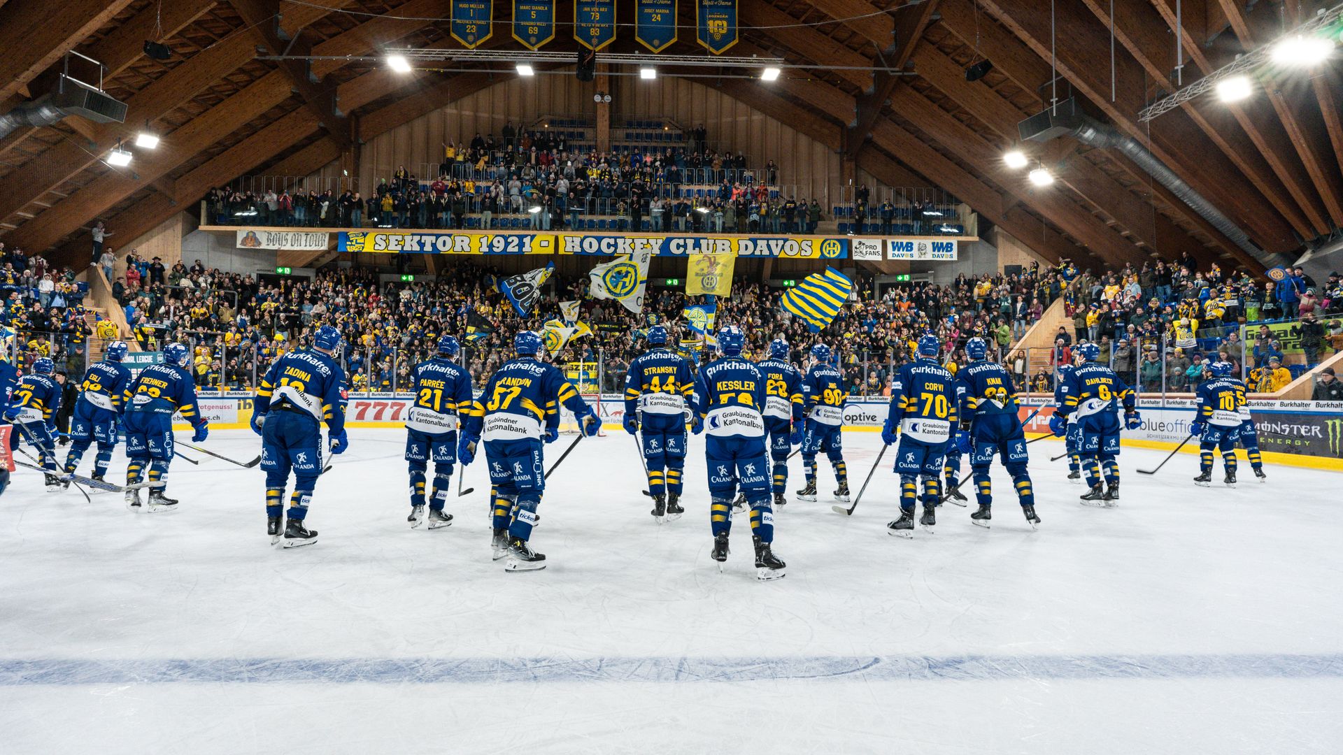 I giocatori dell’HC Davos sono sul ghiaccio dopo la partita e salutano i tifosi, mentre sugli spalti sventolano bandiere gialloblù nello storico stadio del ghiaccio di Davos.
