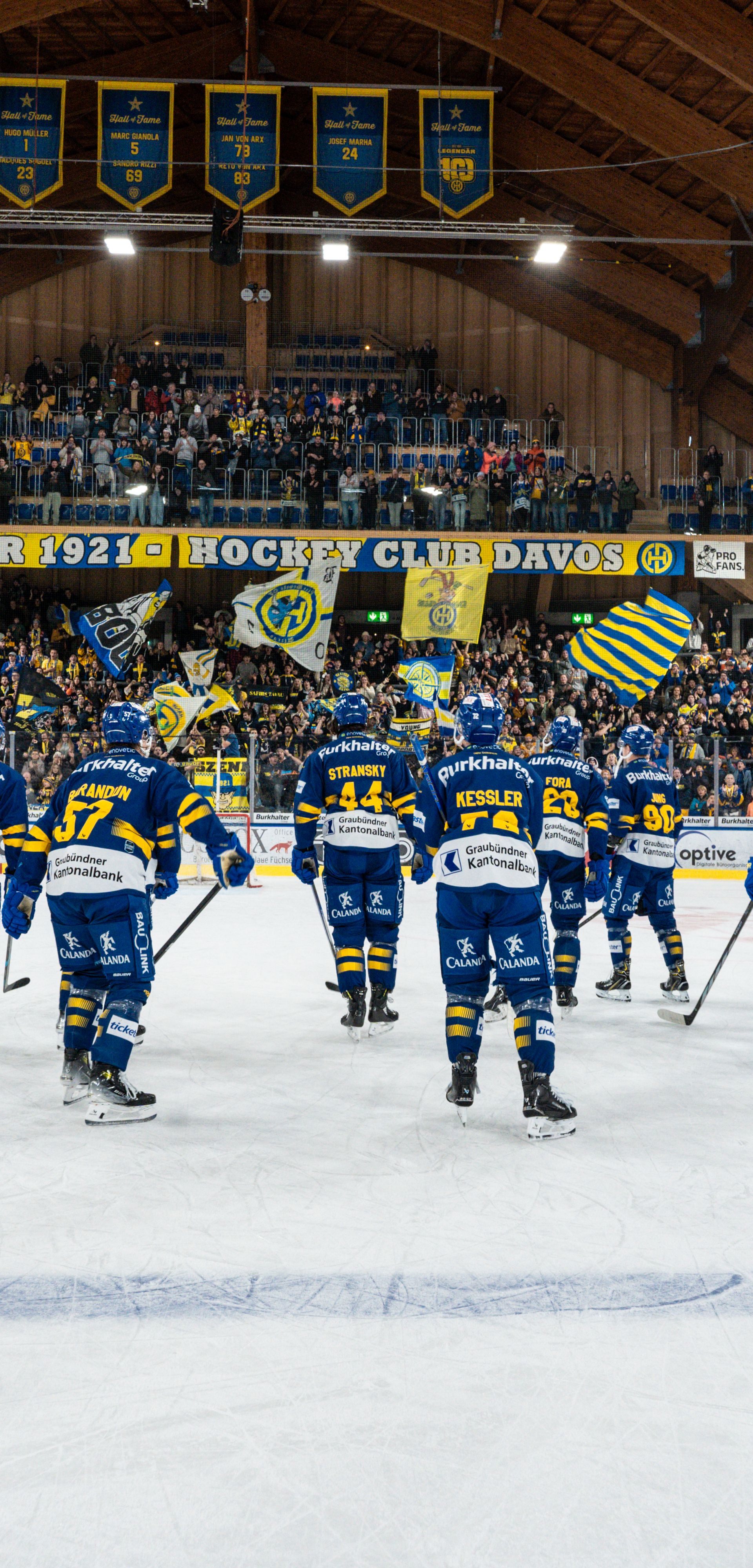 Die Spieler des HC Davos stehen nach dem Spiel auf dem Eis und grüssen ihre Fans. Im Hintergrund jubelt das Publikum mit gelb-blauen Fahnen im legendären Eisstadion Davos.
