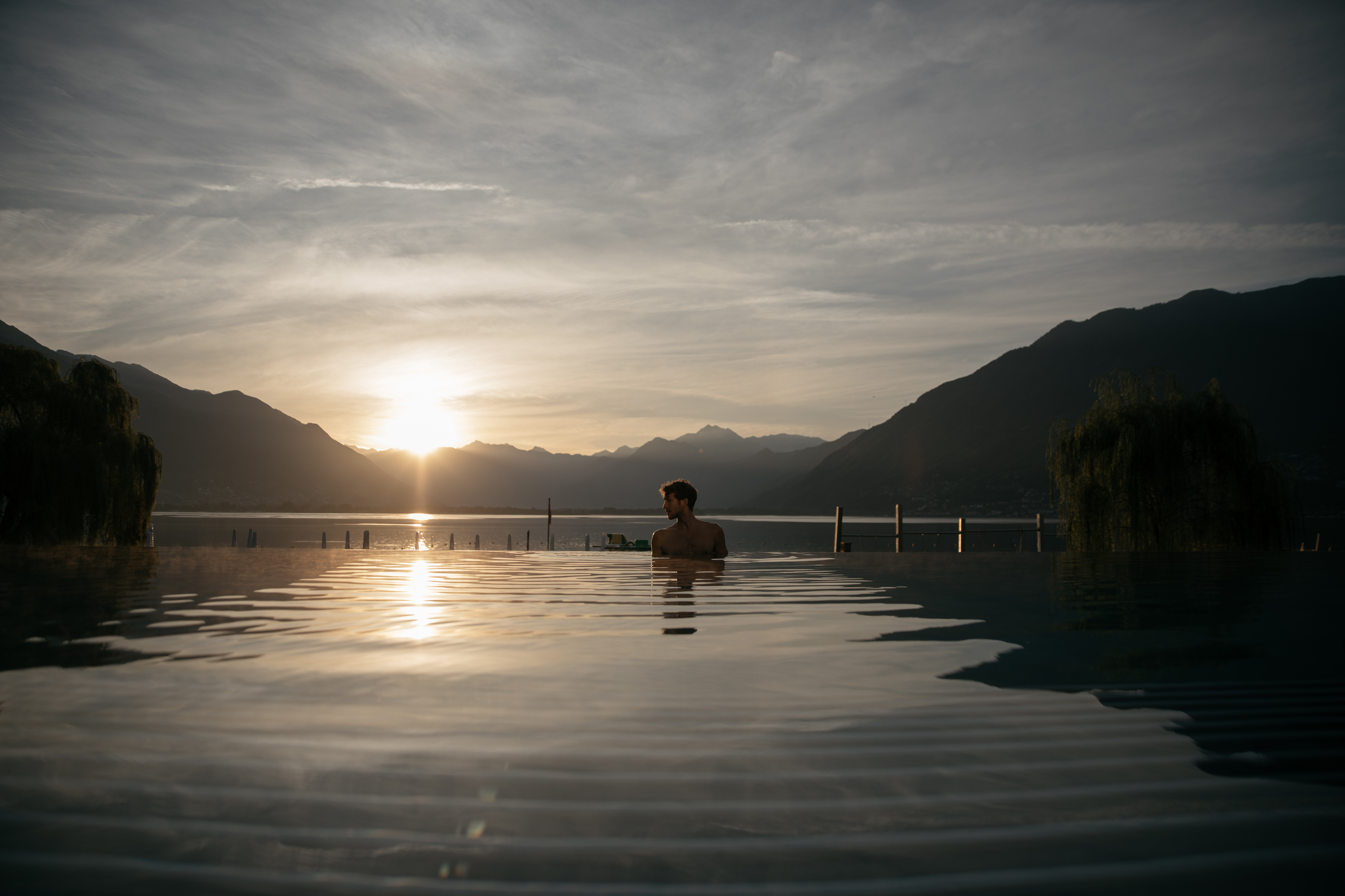 Un uomo si rilassa nella piscina a sfioro delle Termali Salini & Spa Locarno, con vista sul Lago Maggiore al tramonto, circondato da montagne e tranquillità.