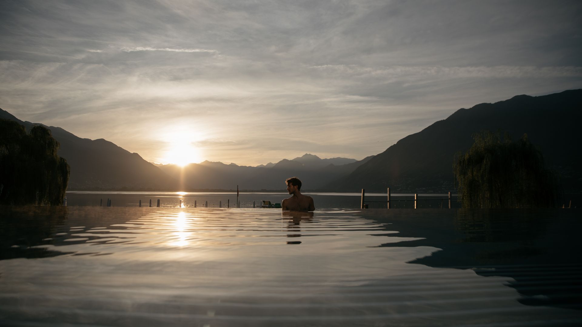 Un uomo si rilassa nella piscina a sfioro delle Termali Salini & Spa Locarno, con vista sul Lago Maggiore al tramonto, circondato da montagne e tranquillità.