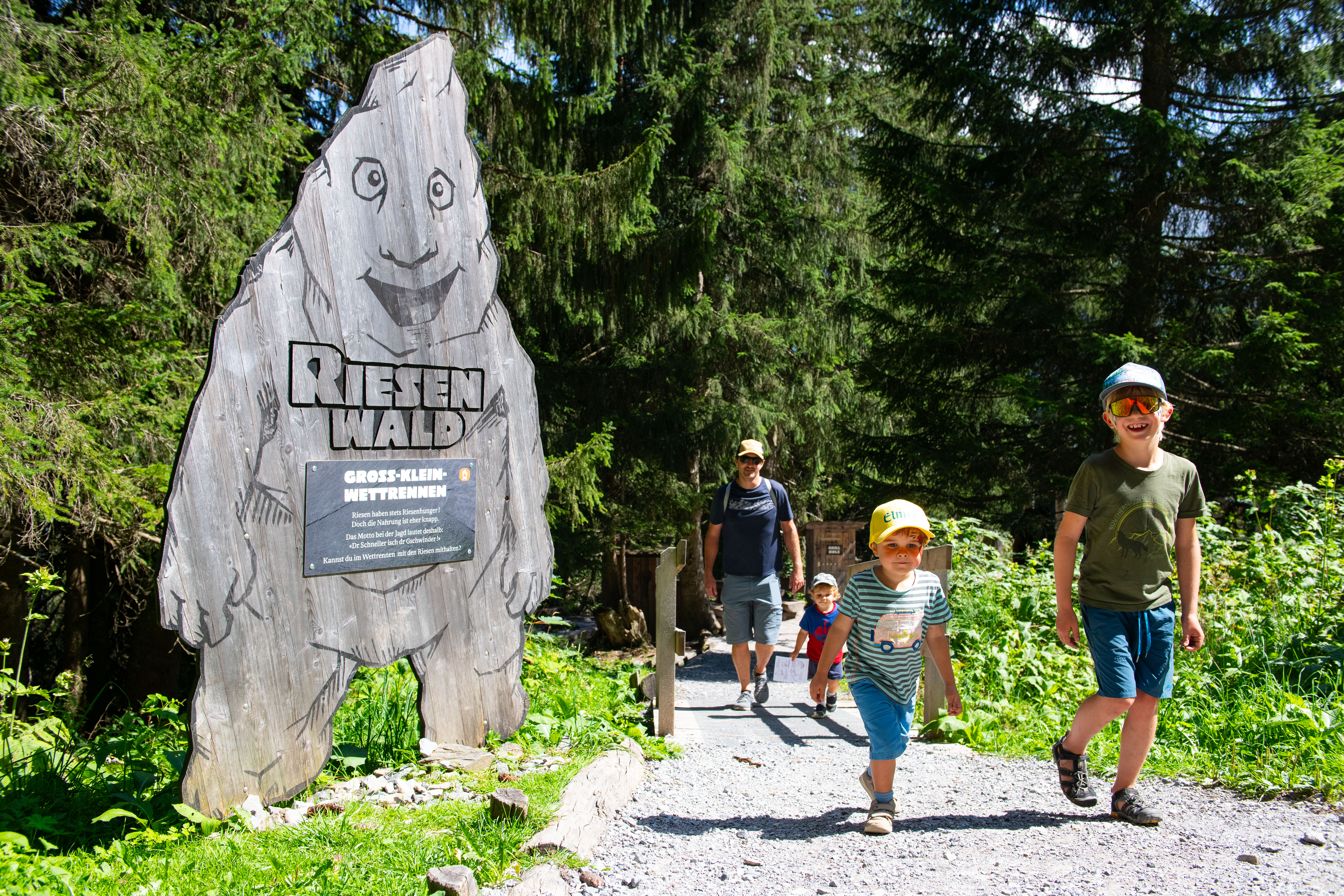 Mehrere Kinder und ein Erwachsener spazieren bei Sonnenschein durch den Riesenwald in Elm – im Vordergrund steht ein hölzernes Riesen-Schild am Waldrand
