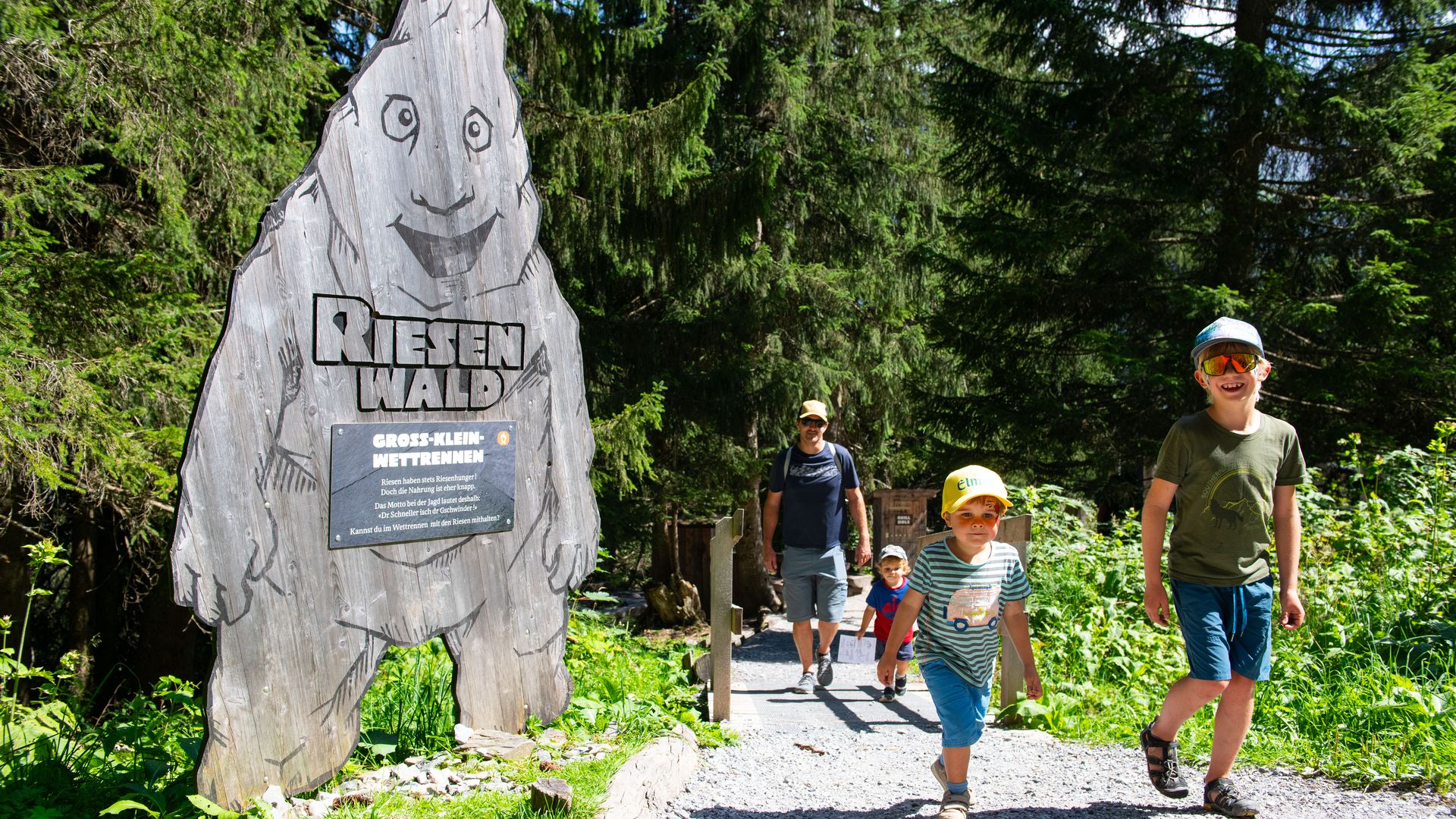 Mehrere Kinder und ein Erwachsener spazieren bei Sonnenschein durch den Riesenwald in Elm – im Vordergrund steht ein hölzernes Riesen-Schild am Waldrand