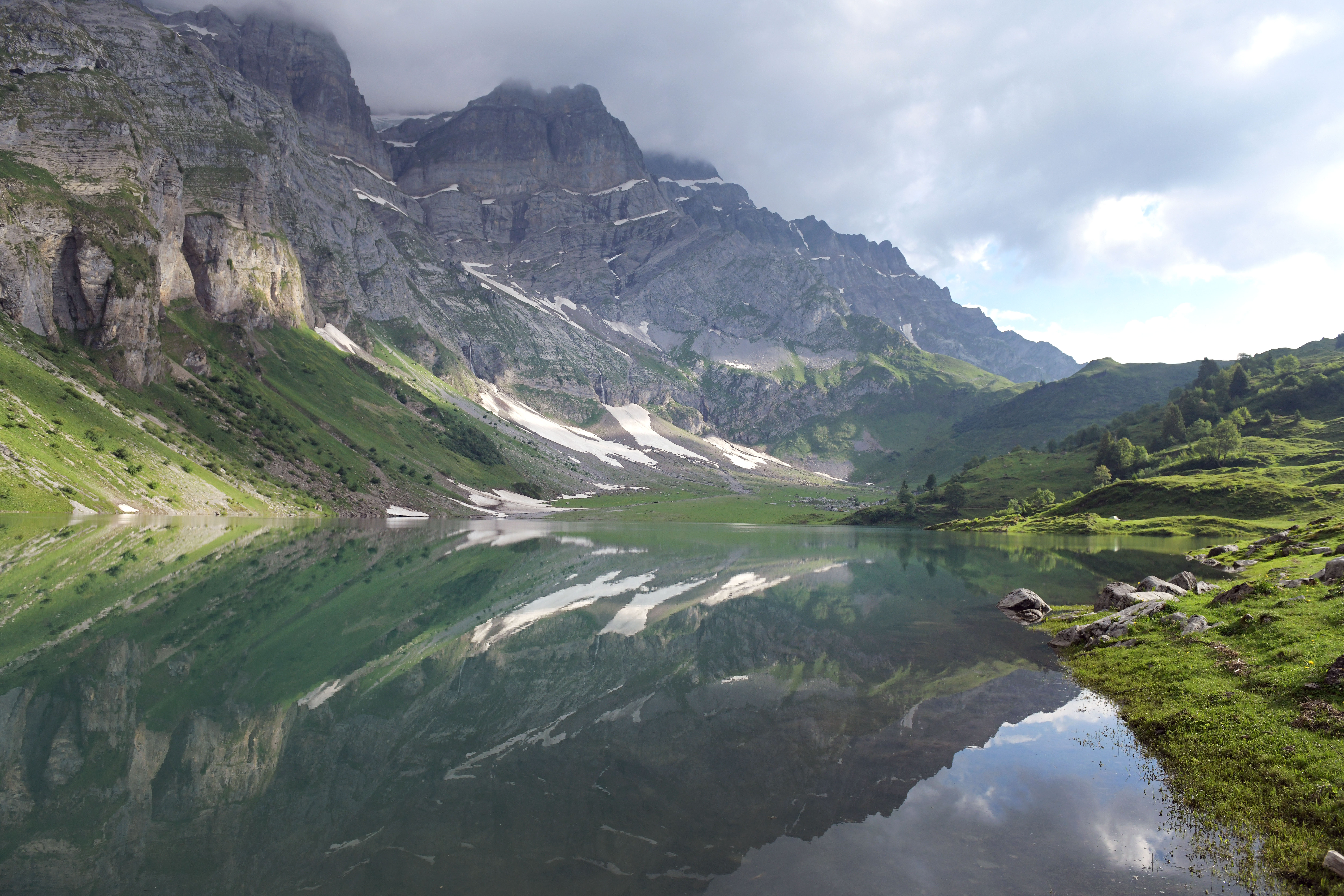  Blick auf den Oberblegisee im Glarnerland: Ein klarer Bergsee spiegelt die umliegenden grünen Hänge und Felswände, darüber ziehen Wolken über die Gipfel.