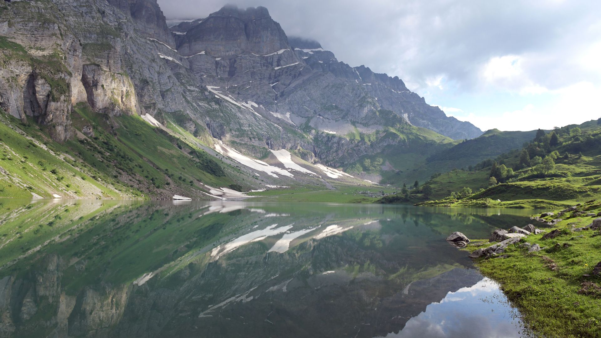 Blick auf den Oberblegisee im Glarnerland: Ein klarer Bergsee spiegelt die umliegenden grünen Hänge und Felswände, darüber ziehen Wolken über die Gipfel.