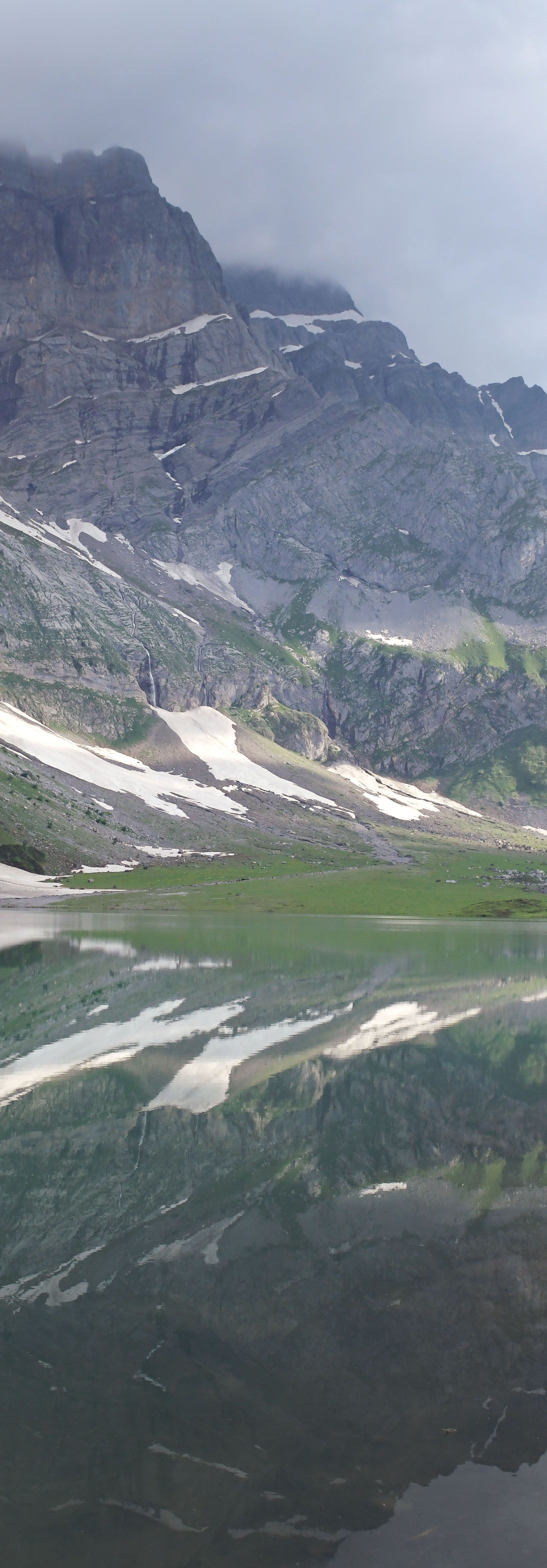 Blick auf den Oberblegisee im Glarnerland: Ein klarer Bergsee spiegelt die umliegenden grünen Hänge und Felswände, darüber ziehen Wolken über die Gipfel.