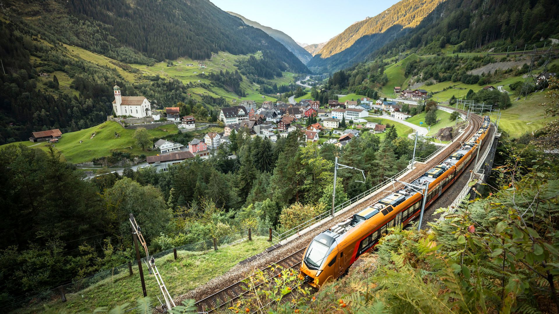 Der kupferfarbene Treno Gottardo fährt auf der historischen Gotthard-Bergstrecke oberhalb von Wassen vorbei, mit Blick auf die ikonische Kirche und das grüne Reusstal im Kanton Uri.